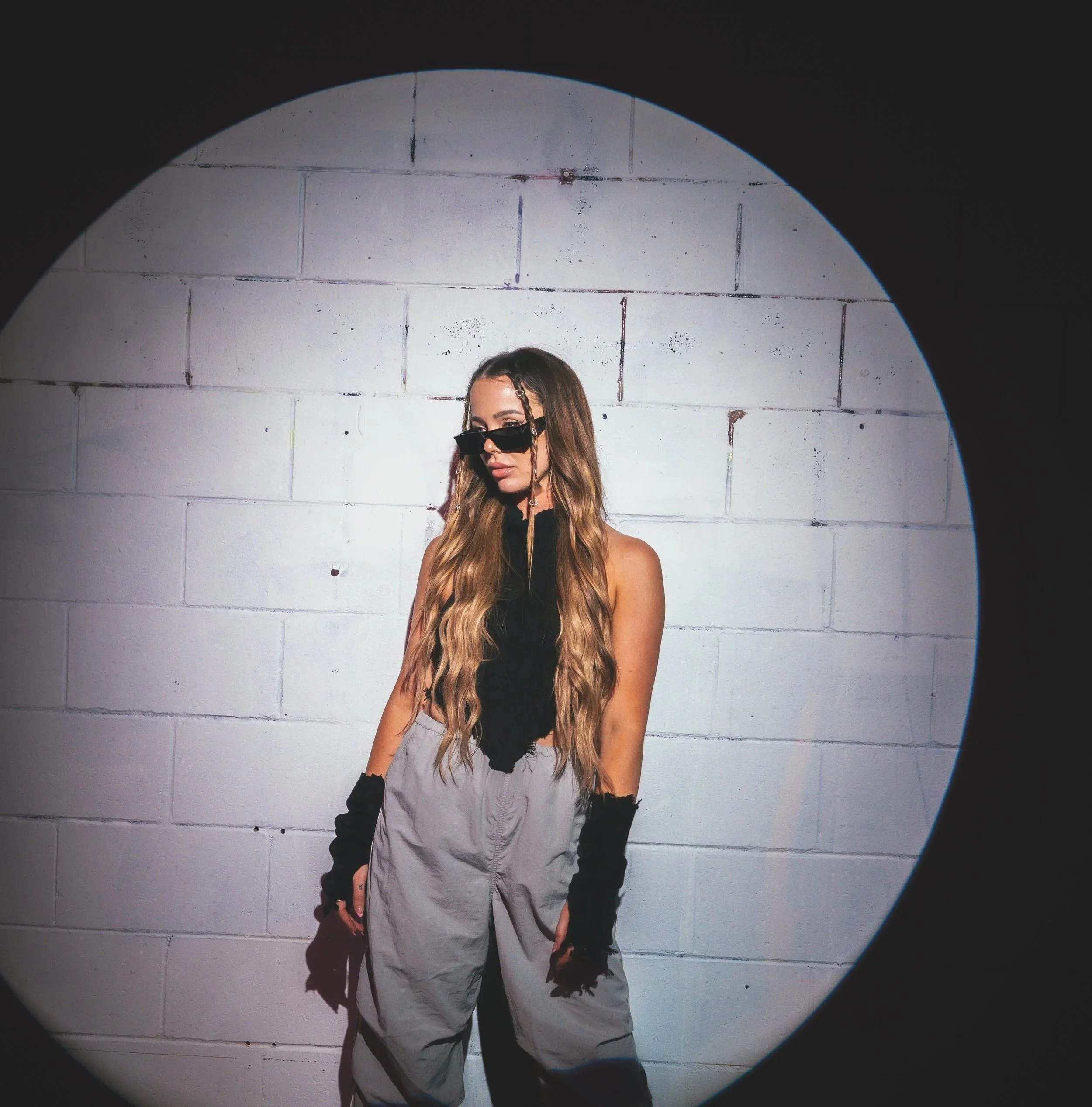 A young woman with long hair wearing sunglasses, a black top, white ripped jeans, and black gloves walking in front of a concrete block wall, illuminated by a circular light source.