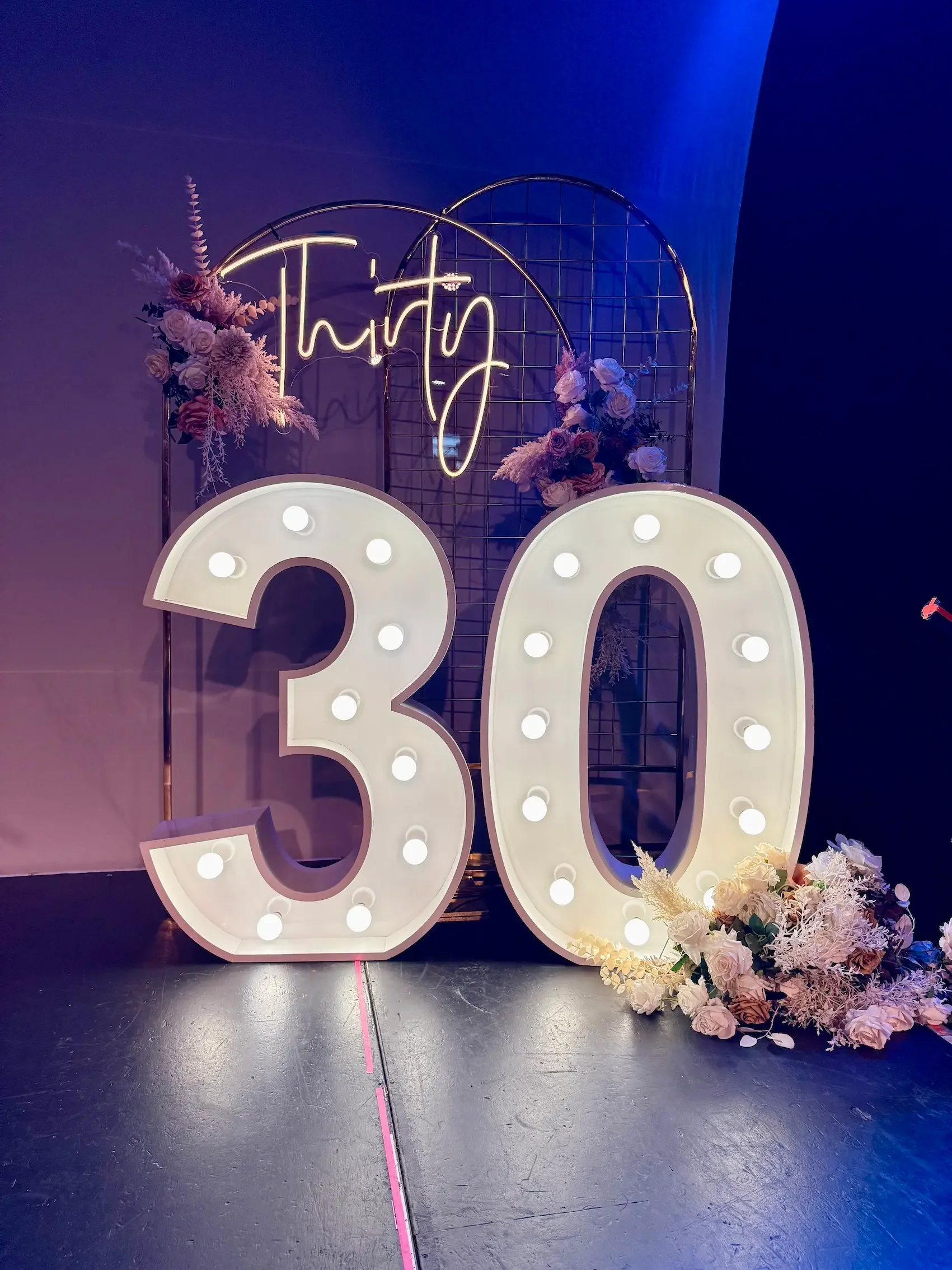 Illuminated wedding or event sign with the word 'happy' in neon lights, decorated with pink and white flowers, and large illuminated numbers '30' on a dark background.