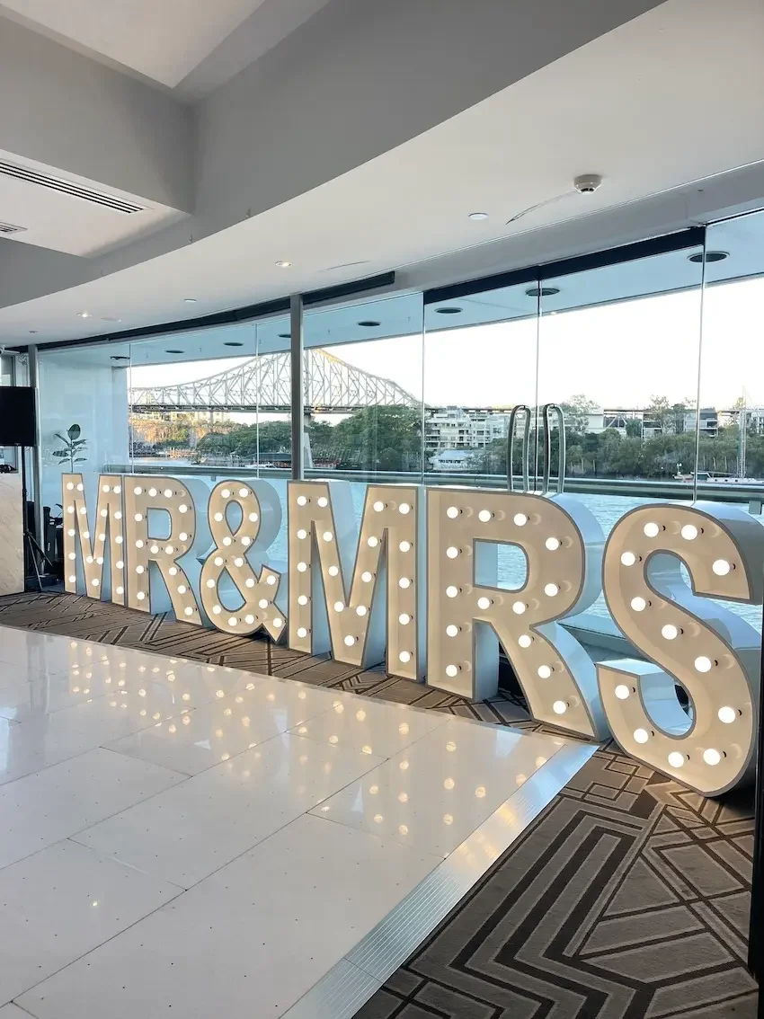 Large illuminated letters spelling "MR & MRS" inside a modern building with floor-to-ceiling windows and a cityscape view in the background.
