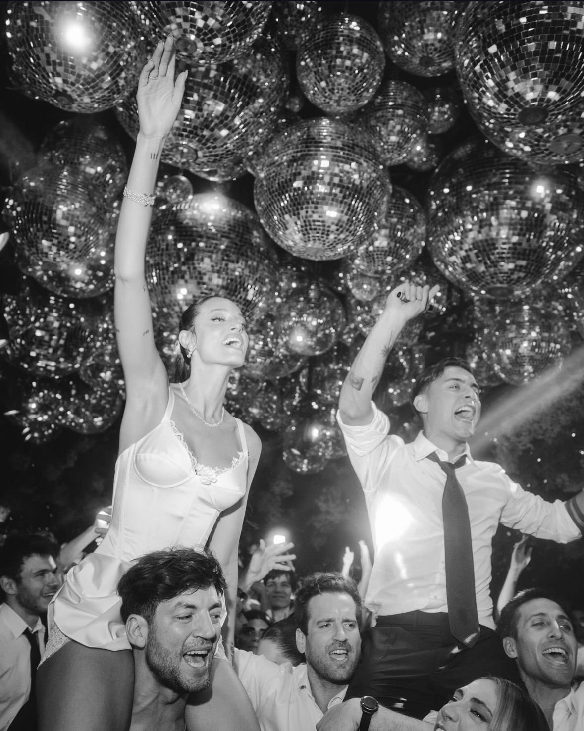 People celebrating at a party with disco balls overhead, some on others' shoulders, enjoying the festive atmosphere.