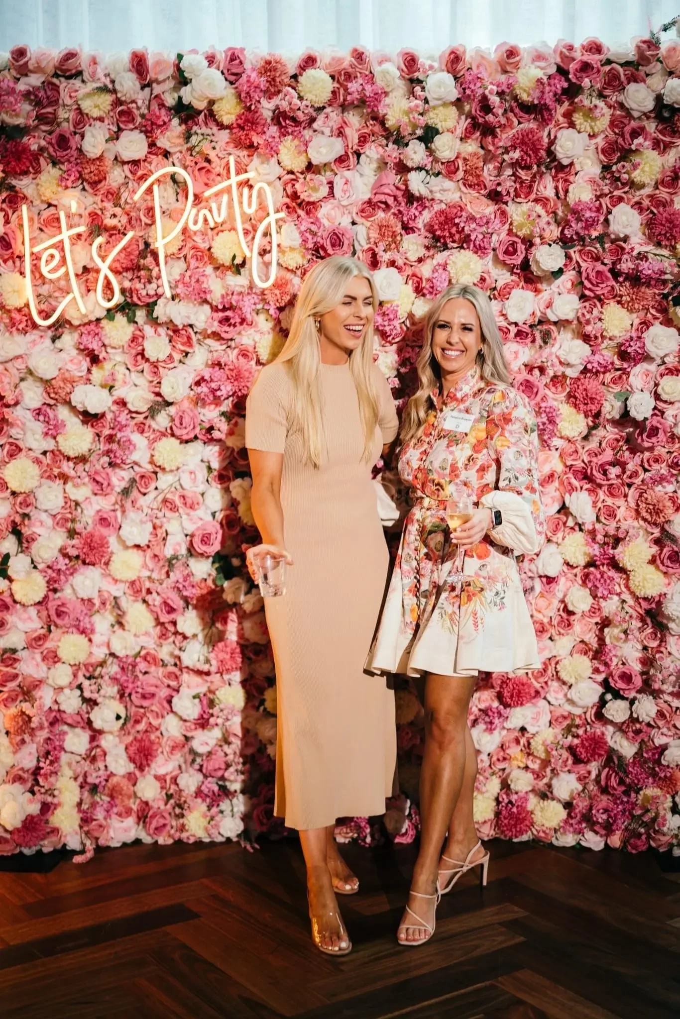 Two women standing in front of a floral wall with a neon sign that says 'Let's Party.' One woman is wearing a beige dress and the other is wearing a floral dress, both smiling and holding drinks.