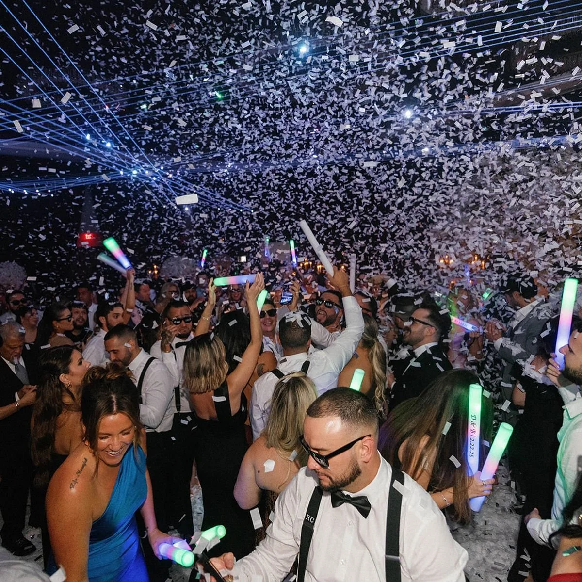 A lively party scene with people dancing, holding glow sticks, and celebrating under a shower of confetti, with laser lights and bright lights illuminating the crowd.