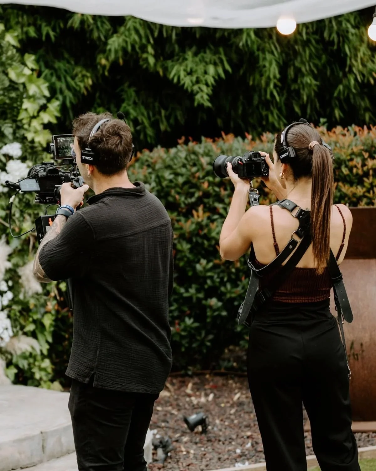 Two people, a man and a woman, wearing headphones and holding cameras with large lenses, filming outdoors against a background of green bushes and trees.