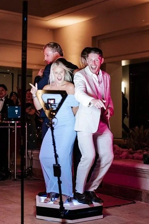 Group of people dancing and having fun at a party, with one woman in a blue dress and two men in suits standing on a small platform, taking a selfie with a camera, in a well-lit indoor setting.