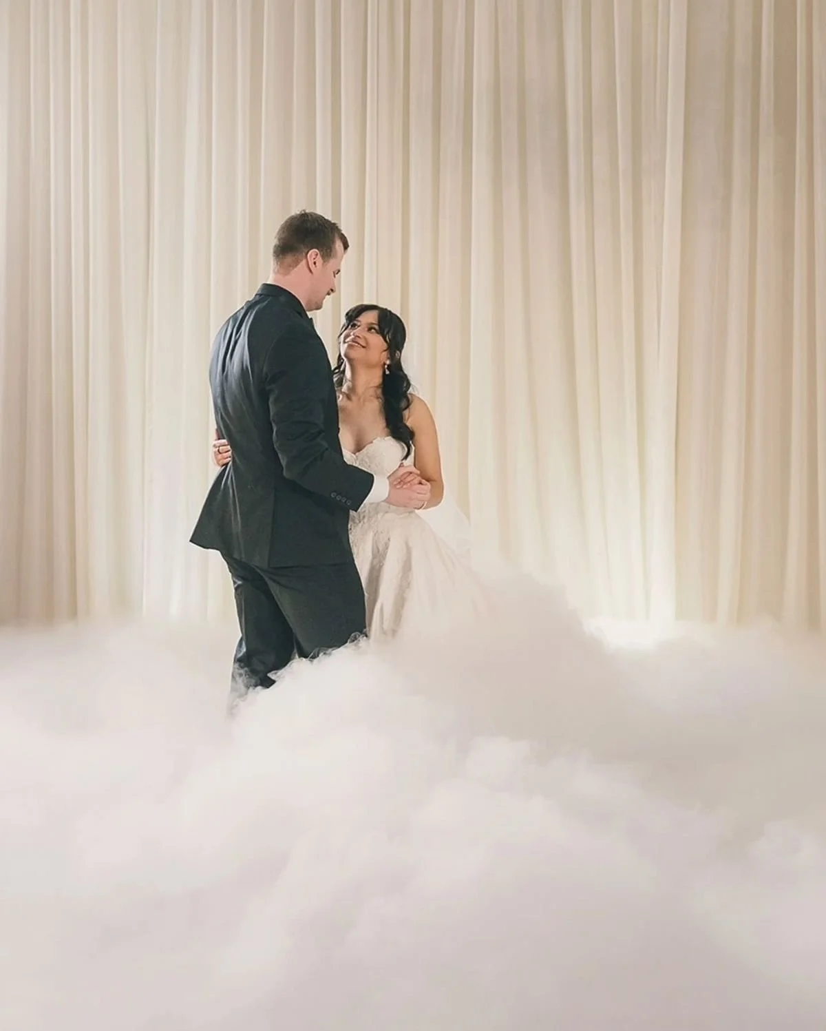 A bride and groom dance together at their wedding reception with a white curtain background and fog on the floor.