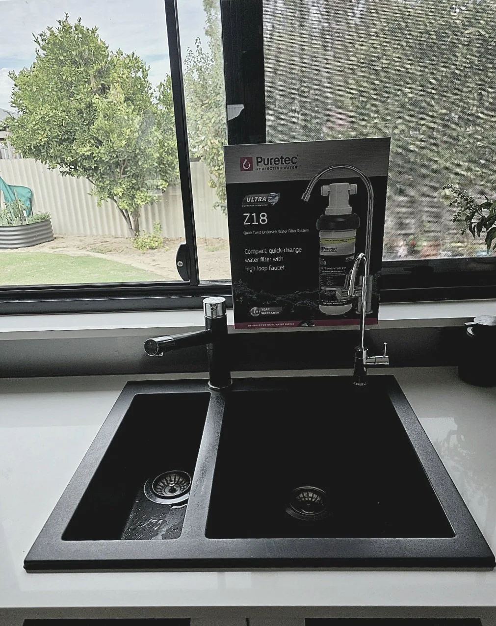 Black kitchen sink with two basins, one with a small amount of water, situated below a window. There is a Puretec water filter system box placed on the sink, indicating it is a Z8 Ultra Undersink Water Filter System. Puretec installer Mandurah.