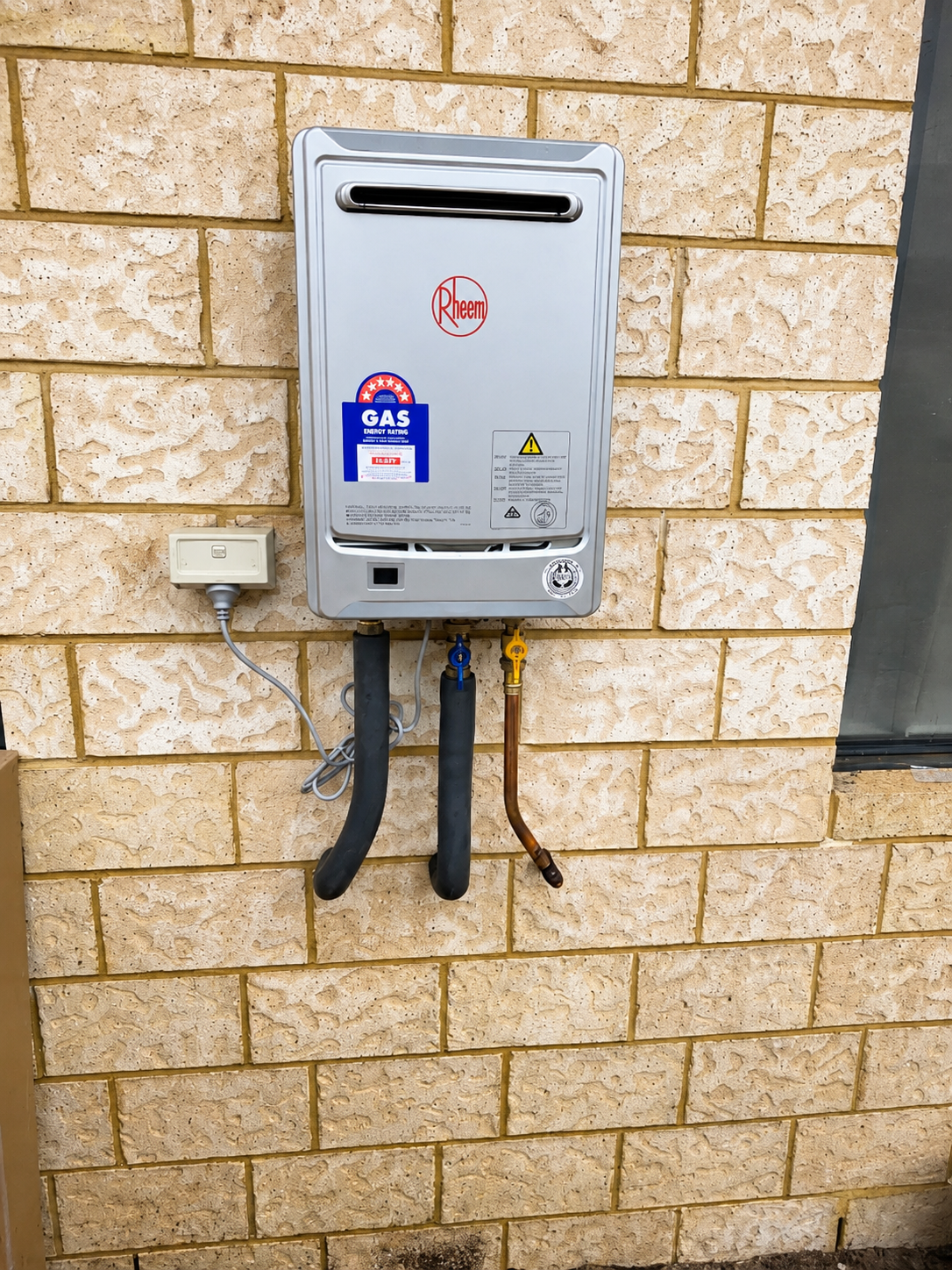 Exterior view of a Rheem gas water heater mounted on a brick wall, with gas and water supply connections, an electrical outlet, and a window nearby.