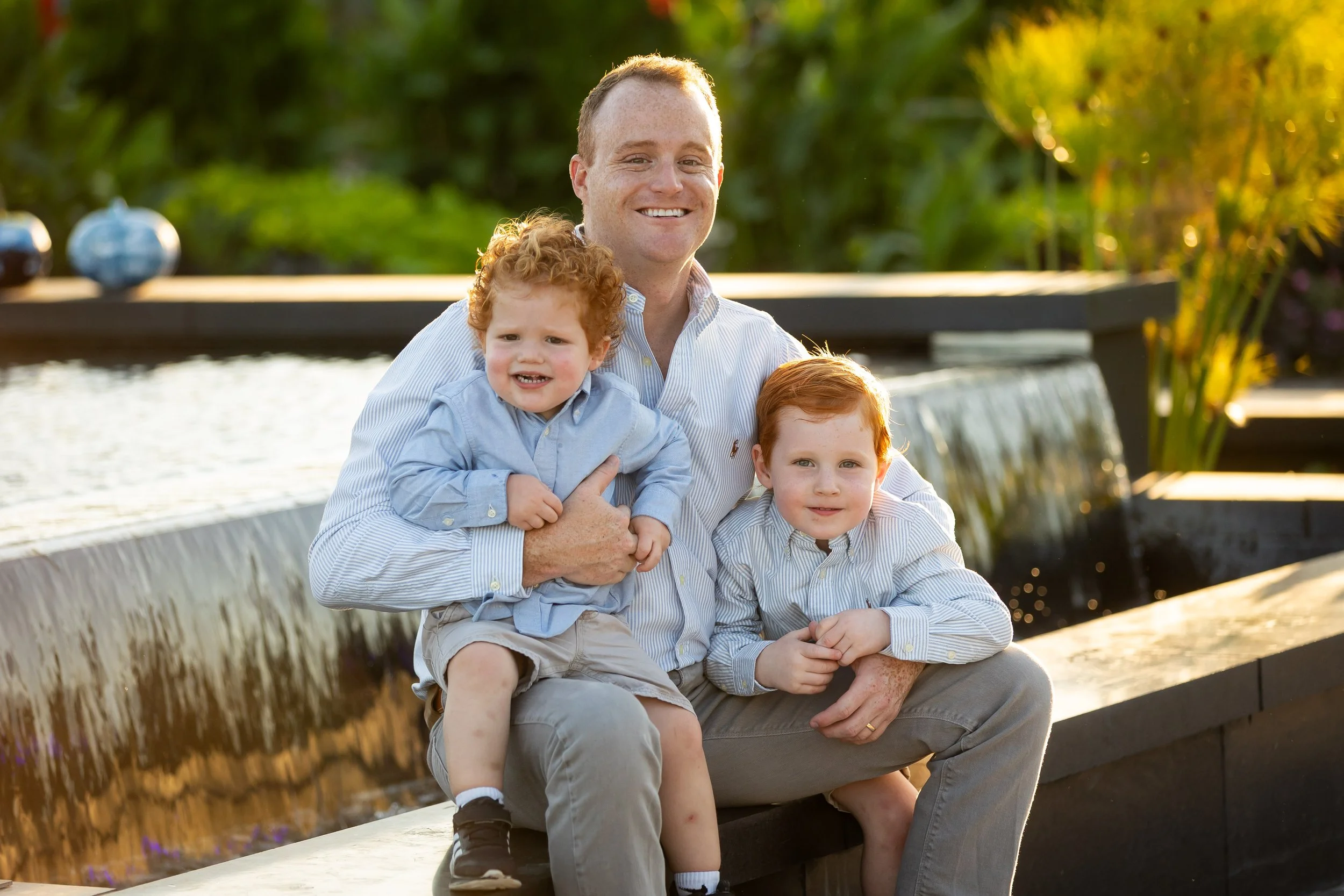 A man sitting on a bench outdoors with three children, all smiling, in a sunny garden with greenery and decorative blue globes in the background.