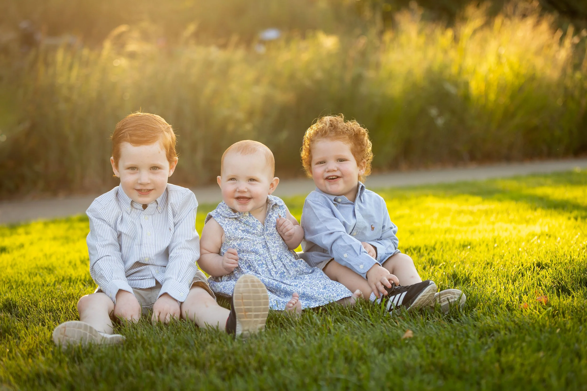 Three young children sitting on grass outdoors, smiling with trees and sunshine in the background.