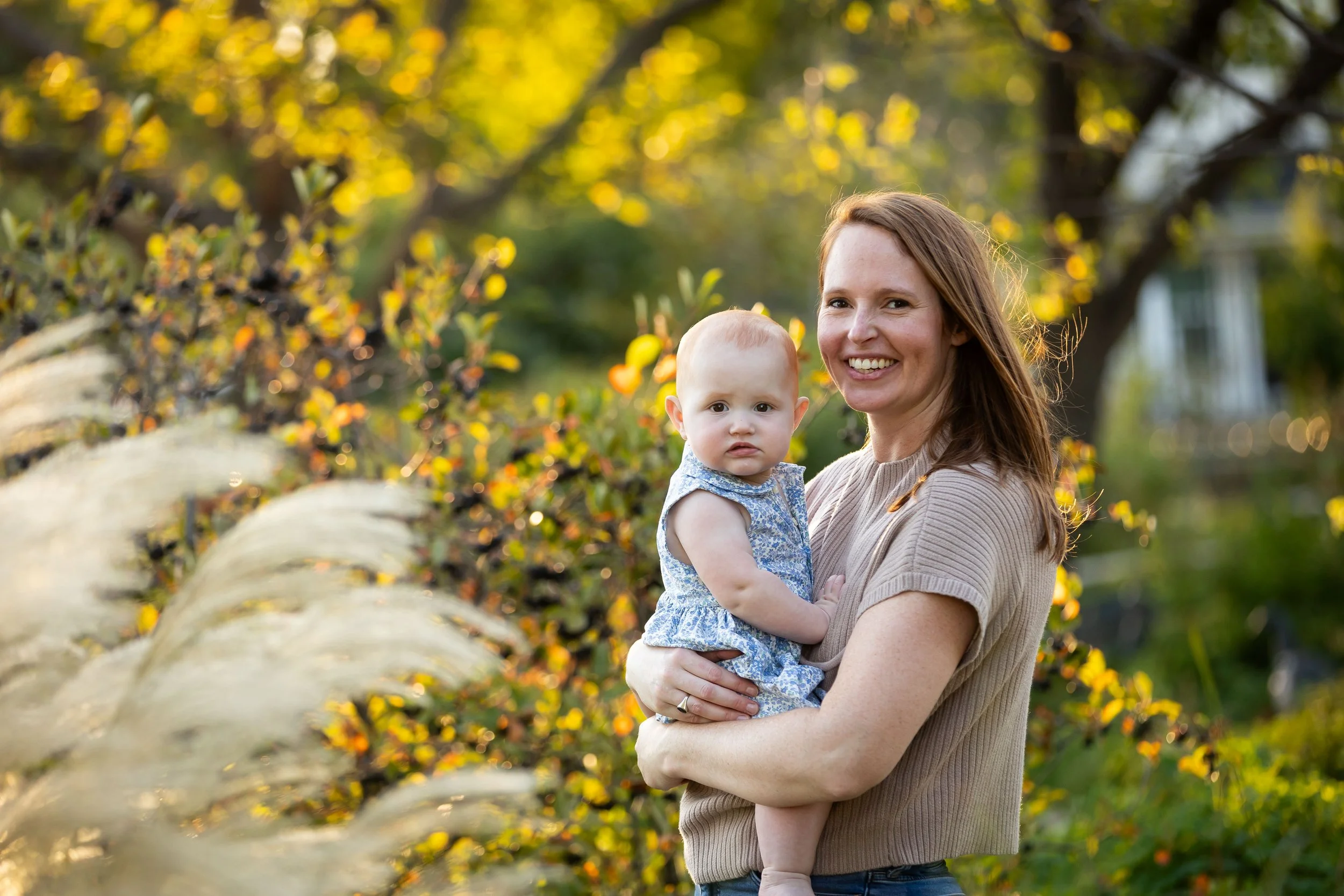 A woman with red hair holding a young girl with light hair, both smiling, outdoors in front of trees with yellow and green leaves, during autumn.