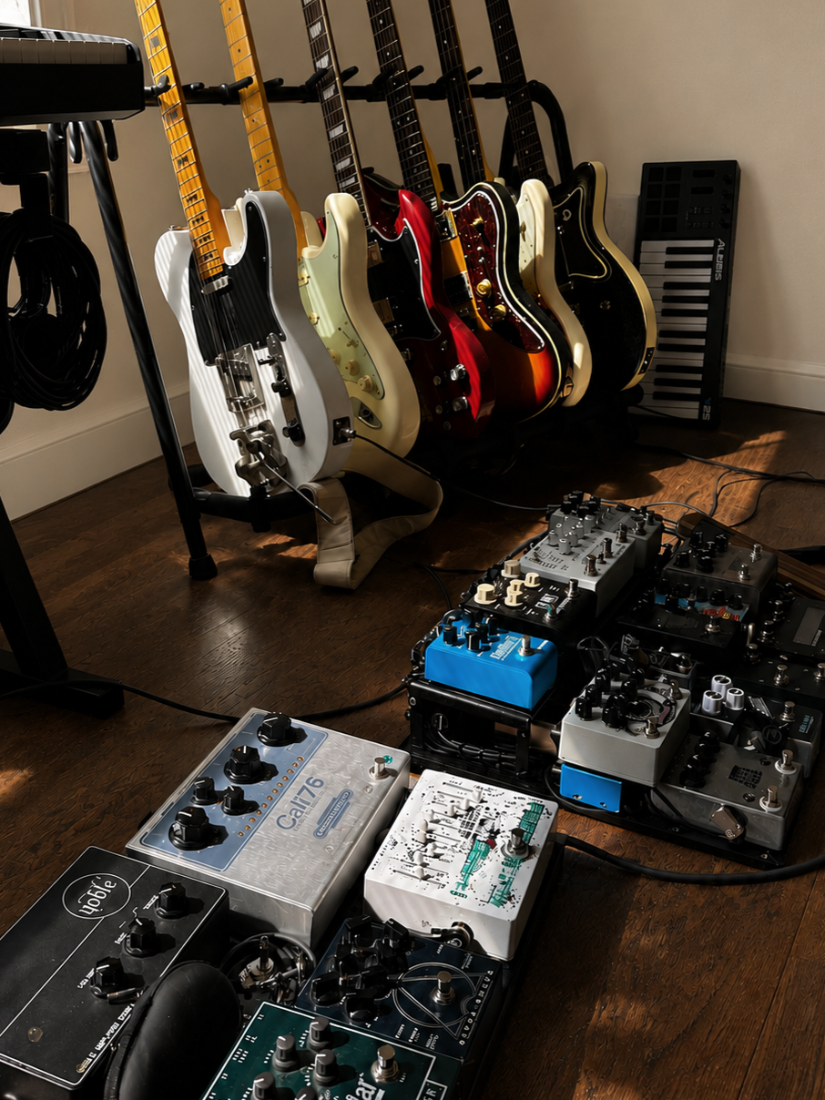 A collection of electric guitars on guitar stands along with various guitar effects pedals and a keyboard in a music studio or practice room.