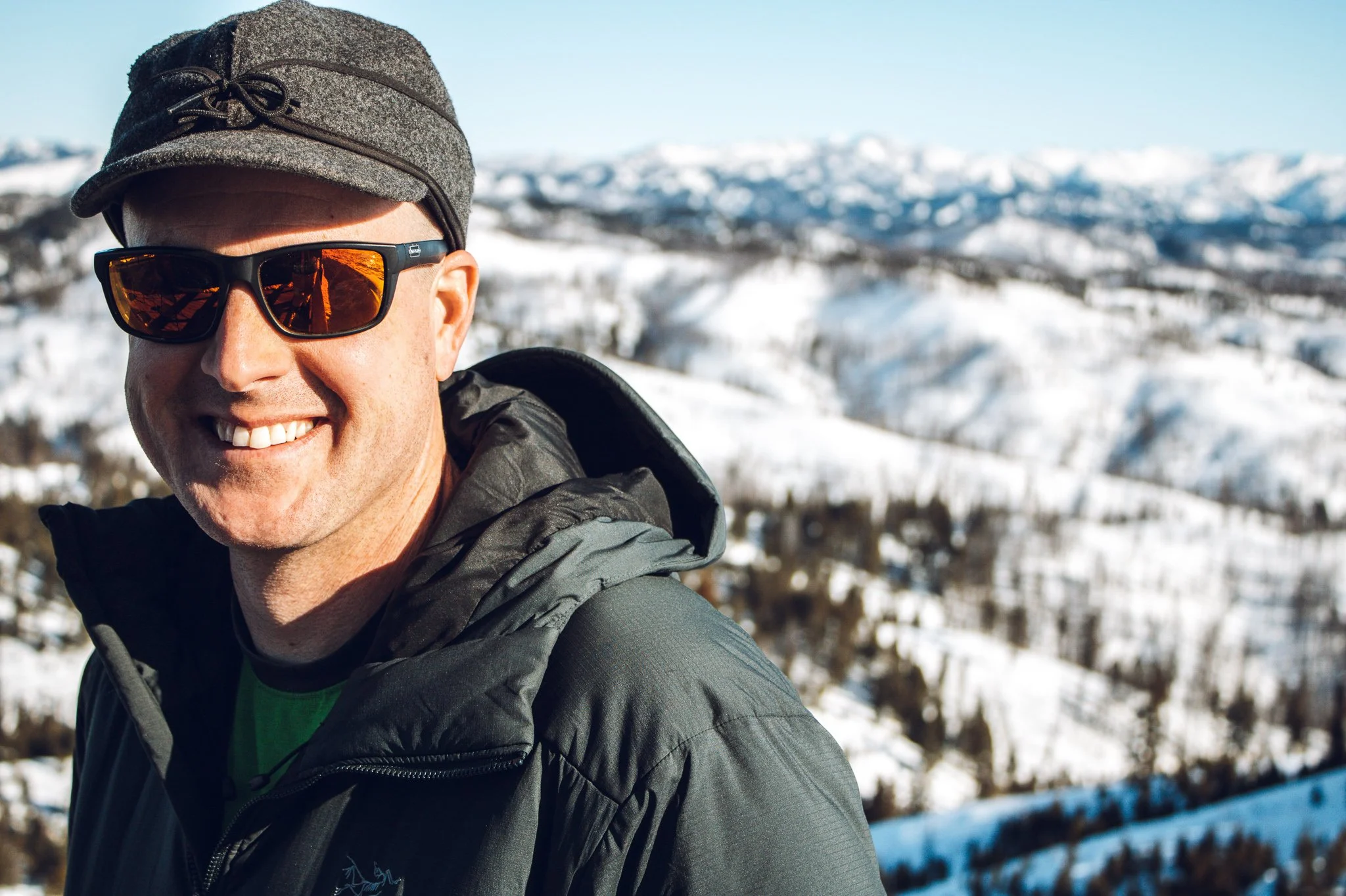 A man wearing sunglasses, a gray jacket, a green shirt, and a black cap smiling outdoors with snow-covered mountains in the background.
