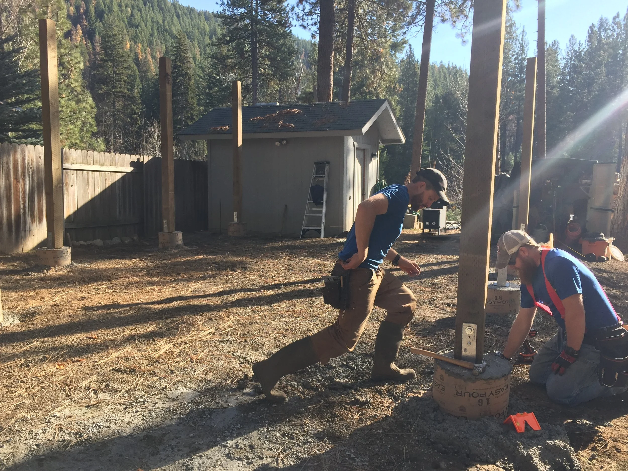 Two men working on construction site outdoors in a wooded area. They are installing supporting beams for a structure, with one man kneeling and the other bending over. There is a small shed in the background, along with tall trees and a mountain landscape.