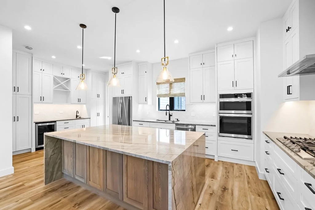 Bright modern kitchen with white cabinets, a large marble-topped island, stainless steel appliances, hardwood floors, and pendant lighting.
