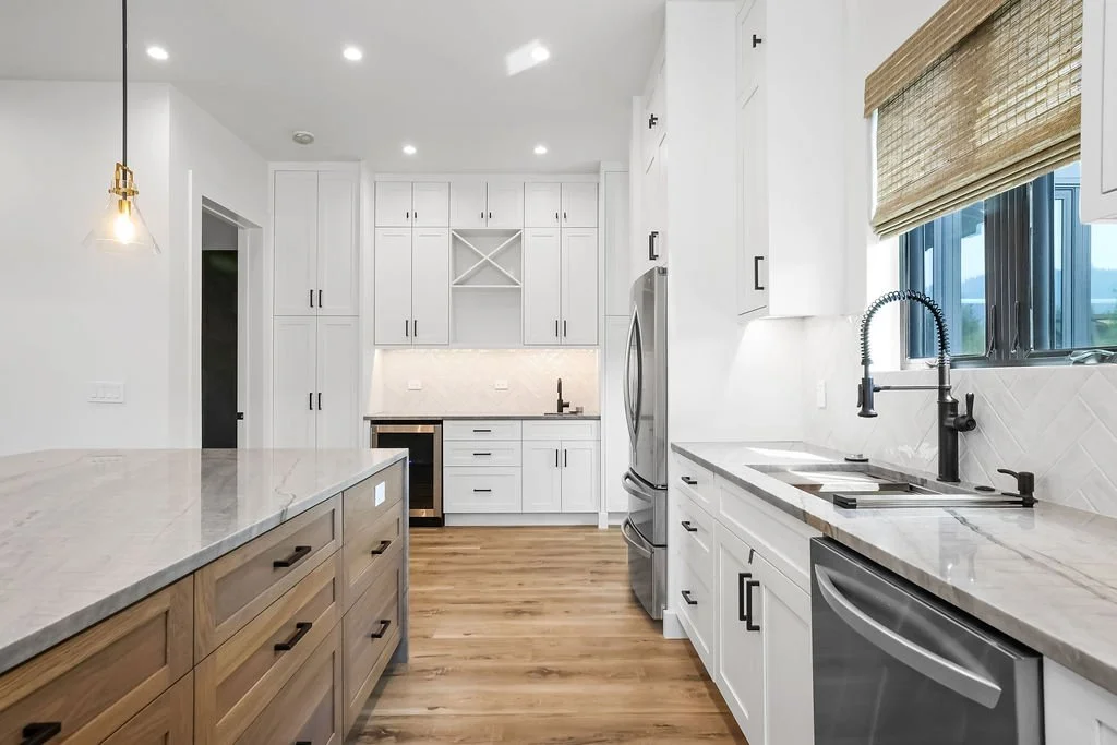 Modern white kitchen with wooden floors, black handles, stainless steel refrigerator, and large window with bamboo blind.