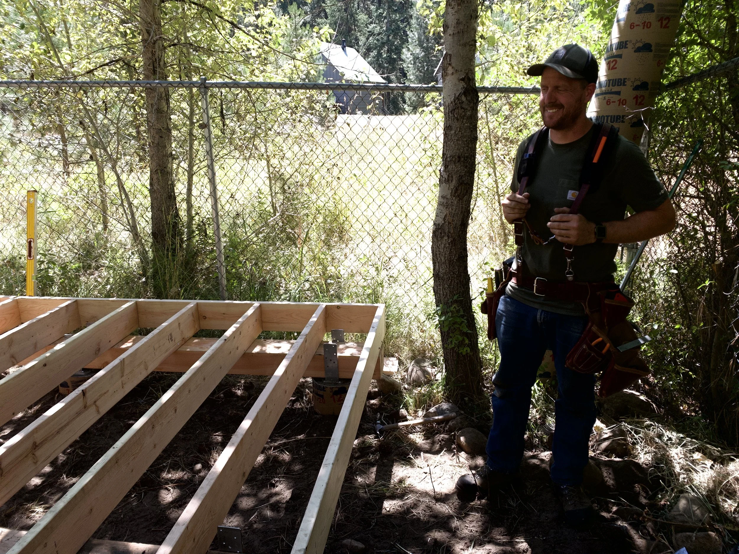 Man smiling, wearing a cap and backpack, standing outdoors near a chain-link fence and a tree, with construction framing visible in the foreground, surrounded by trees and foliage.