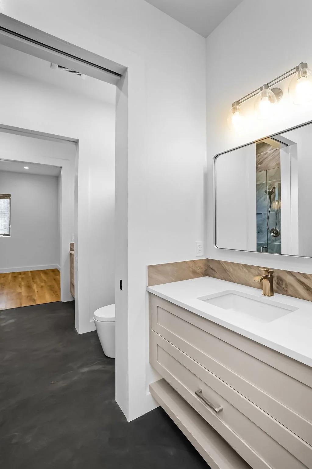 Modern bathroom with white walls, beige vanity, gold faucet, large mirror, and a visible shower in the reflection. Part of the adjacent room with wood flooring is also visible.