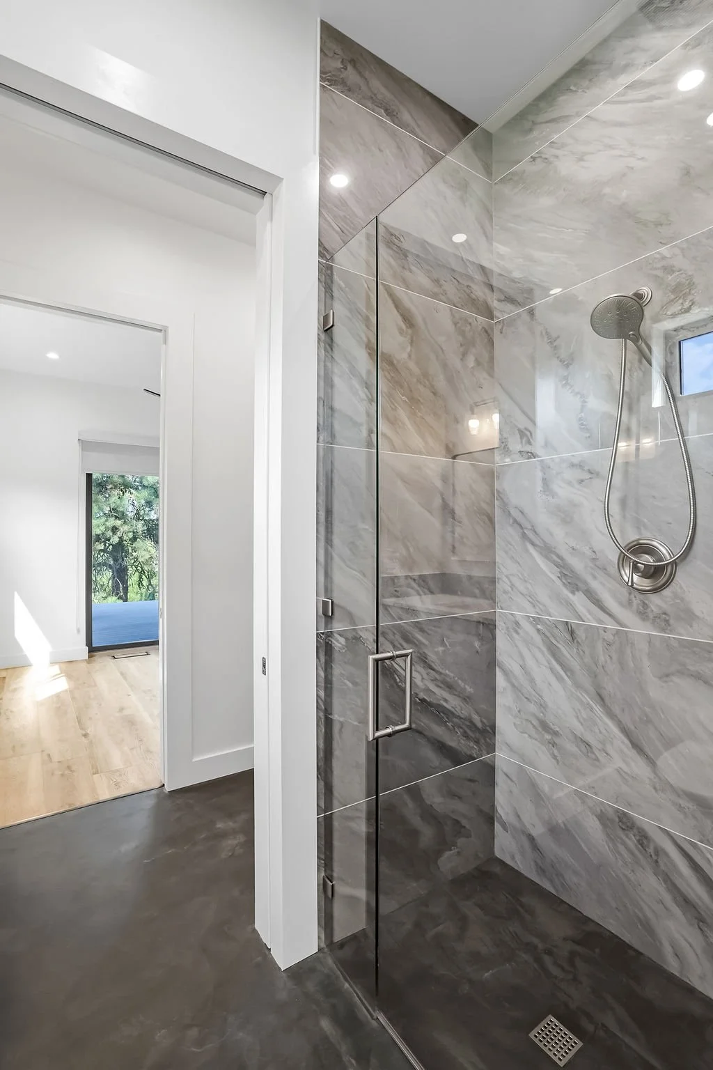 Modern walk-in shower with gray marble tiles, glass door, and handheld showerhead in a bright bathroom.