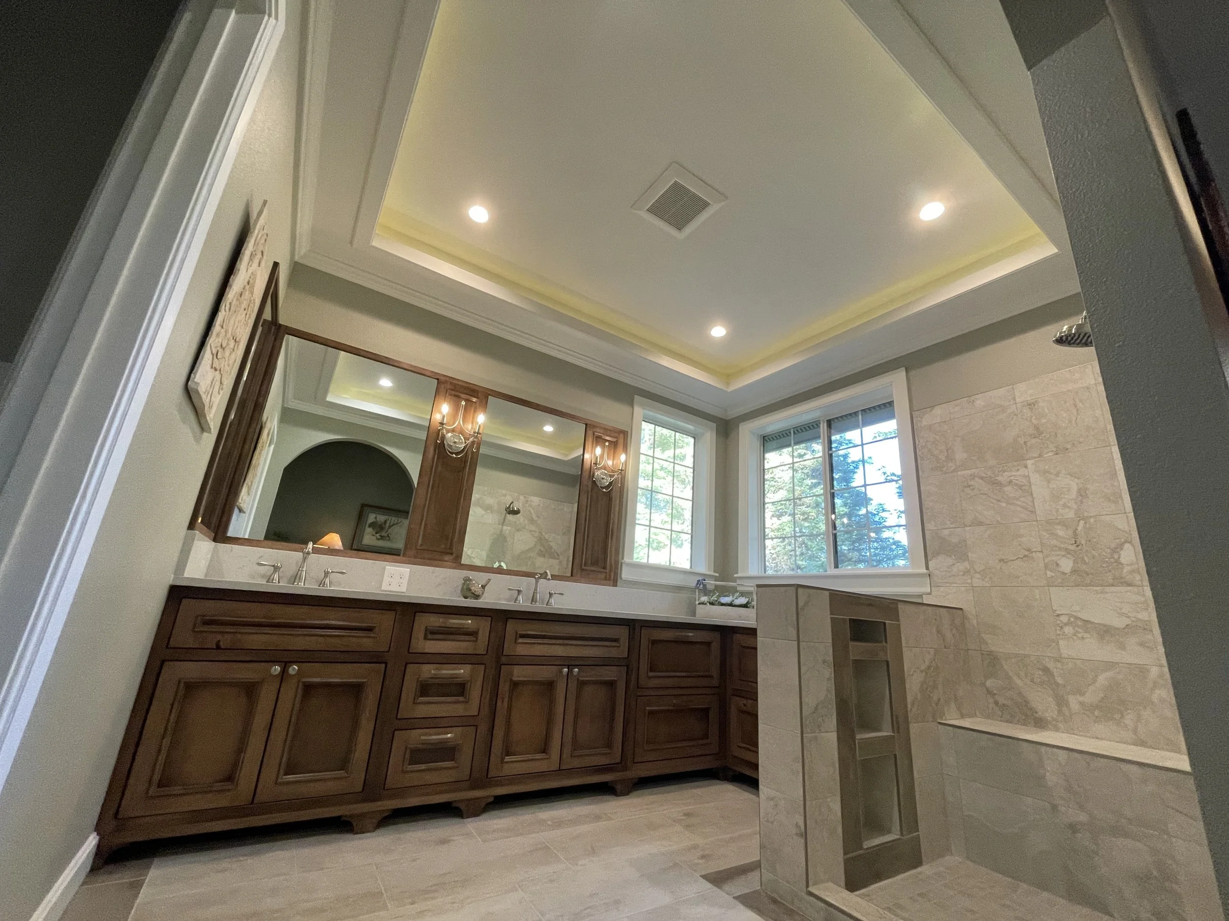 Bathroom with a double vanity with dark wood cabinets, a large mirror, granite countertop, and a sink. The bathroom has a window, tile walls, and a shower enclosure with a tiled interior.
