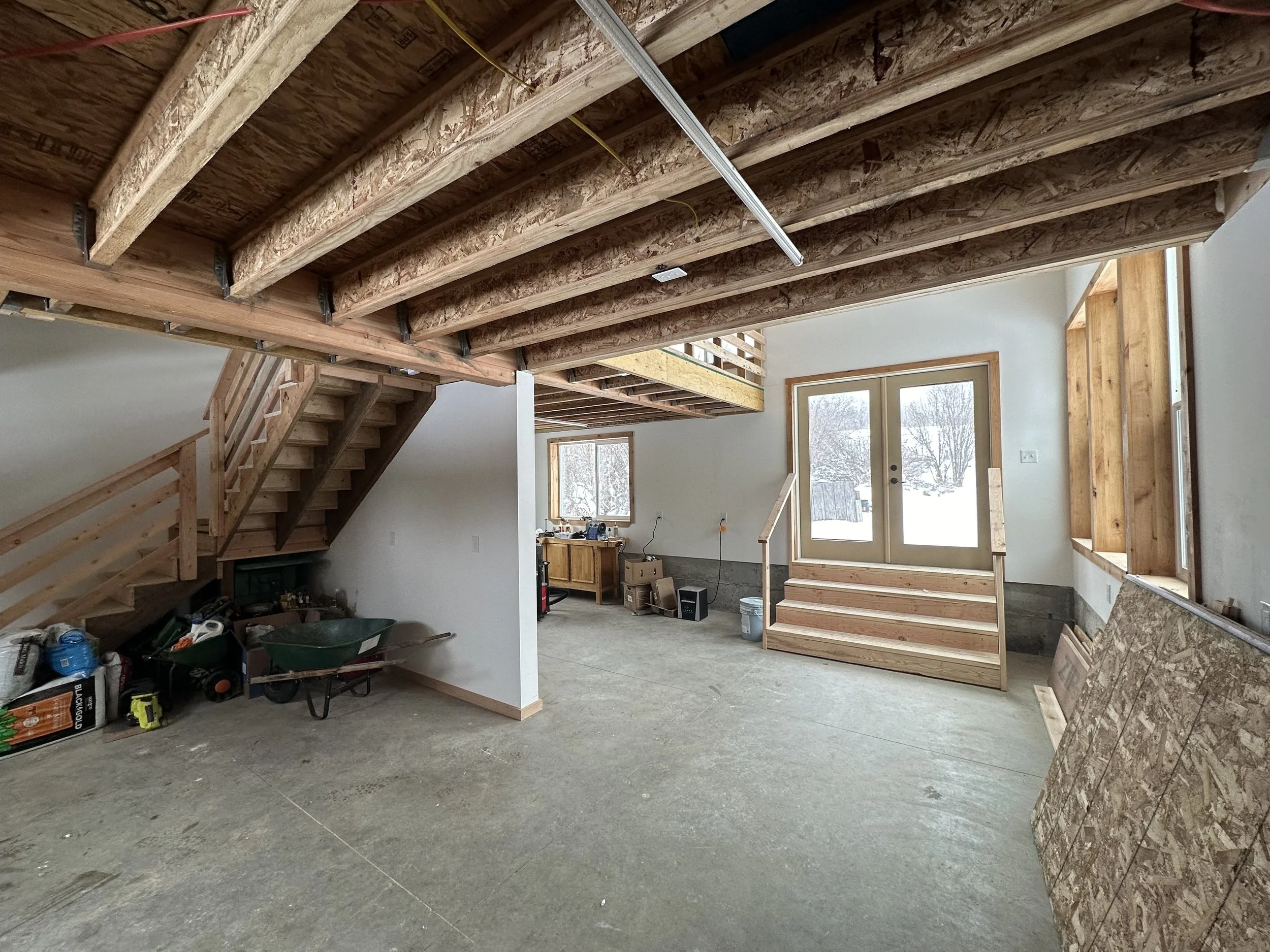 Interior of a house under construction. Exposed ceiling beams, staircase, window, and unfinished walls with tools and materials on the floor.