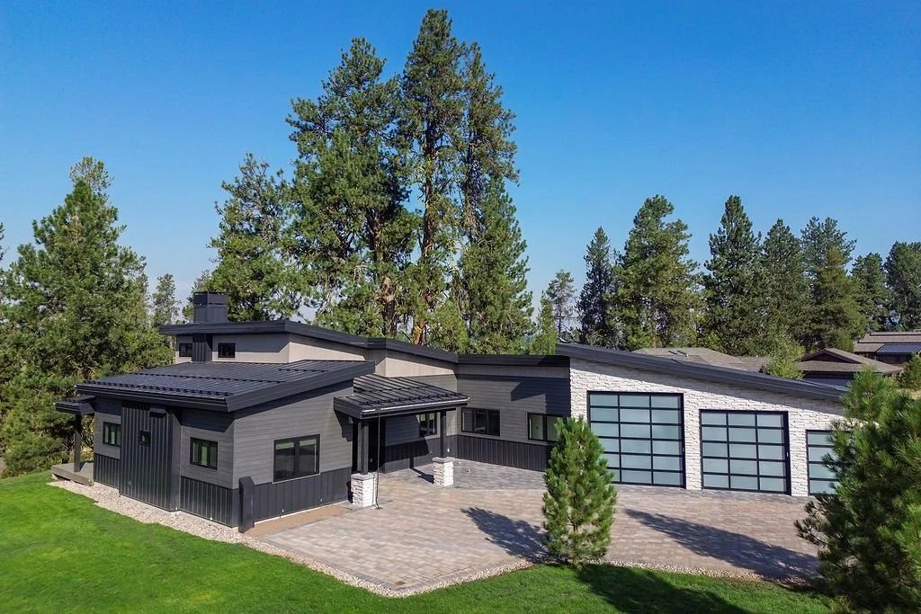 Modern house with dark gray exterior, large glass garage doors, and surrounding trees, situated on a lawn with green grass and a paved driveway.