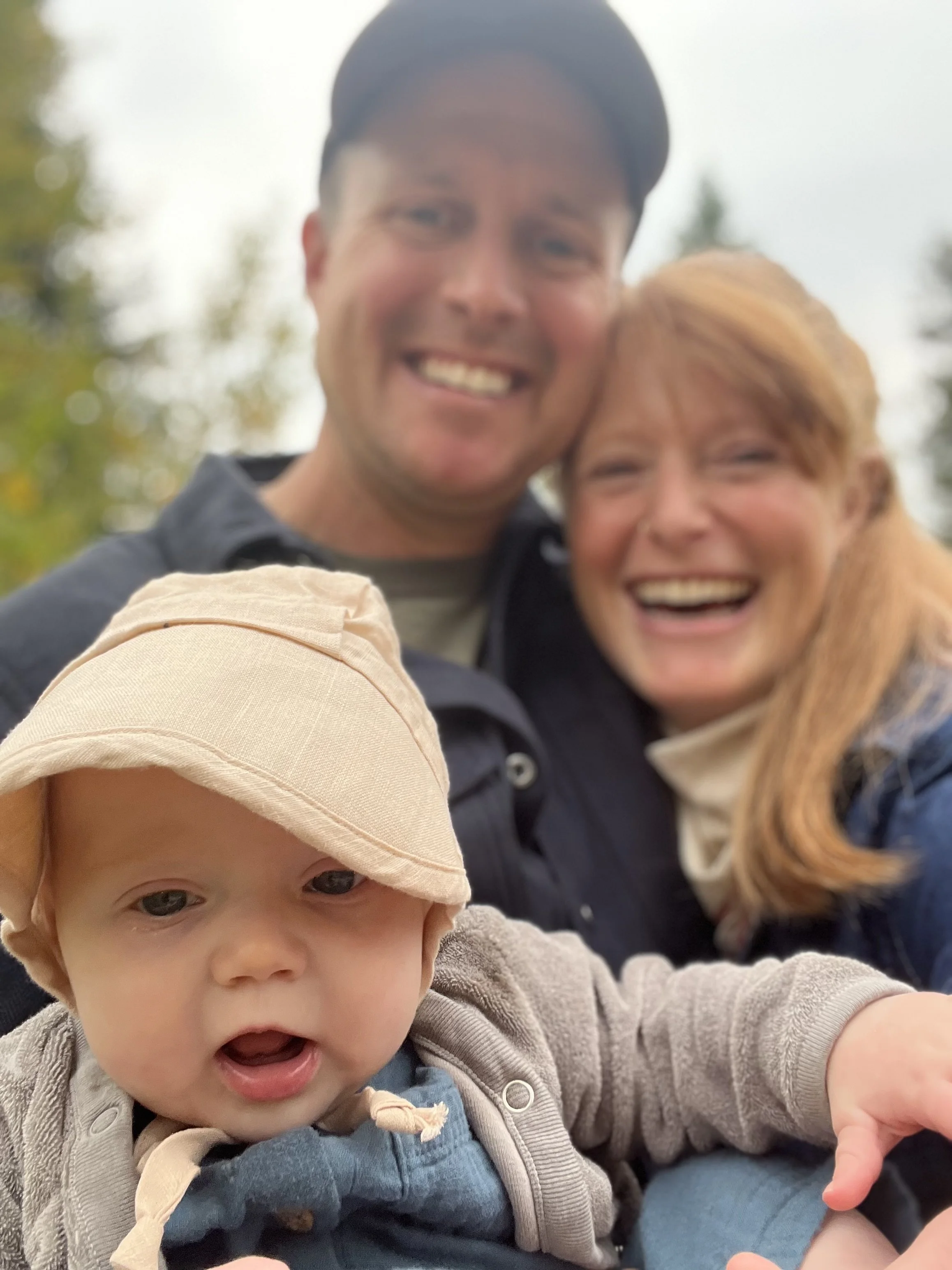 A smiling man and woman taking a selfie outdoors with a young child in the foreground, wearing a beige hat and looking at the camera.