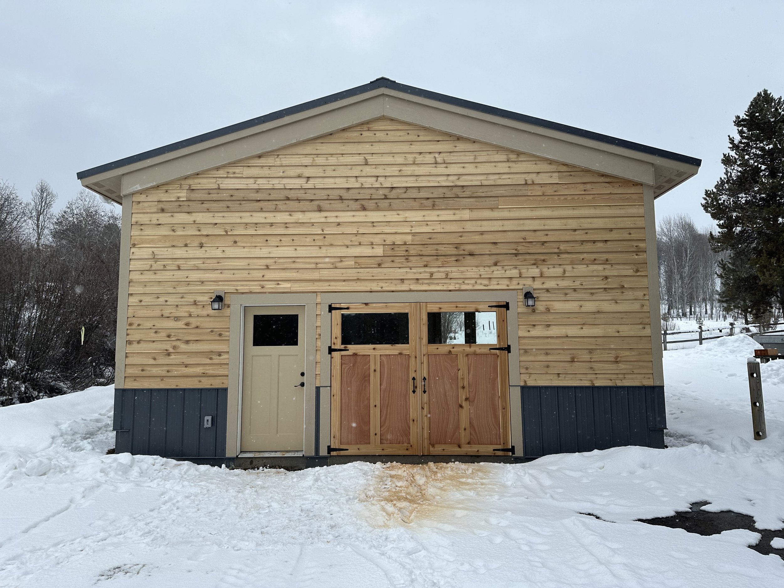 A small wooden shed with a gable roof in a snowy landscape, with trees and overcast sky in the background.