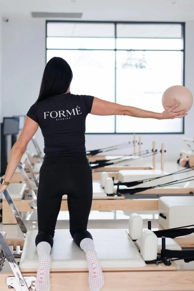 A woman in athletic clothing performing a Pilates exercise on a reformer machine in a fitness studio, holding a small exercise ball with her extended arm.