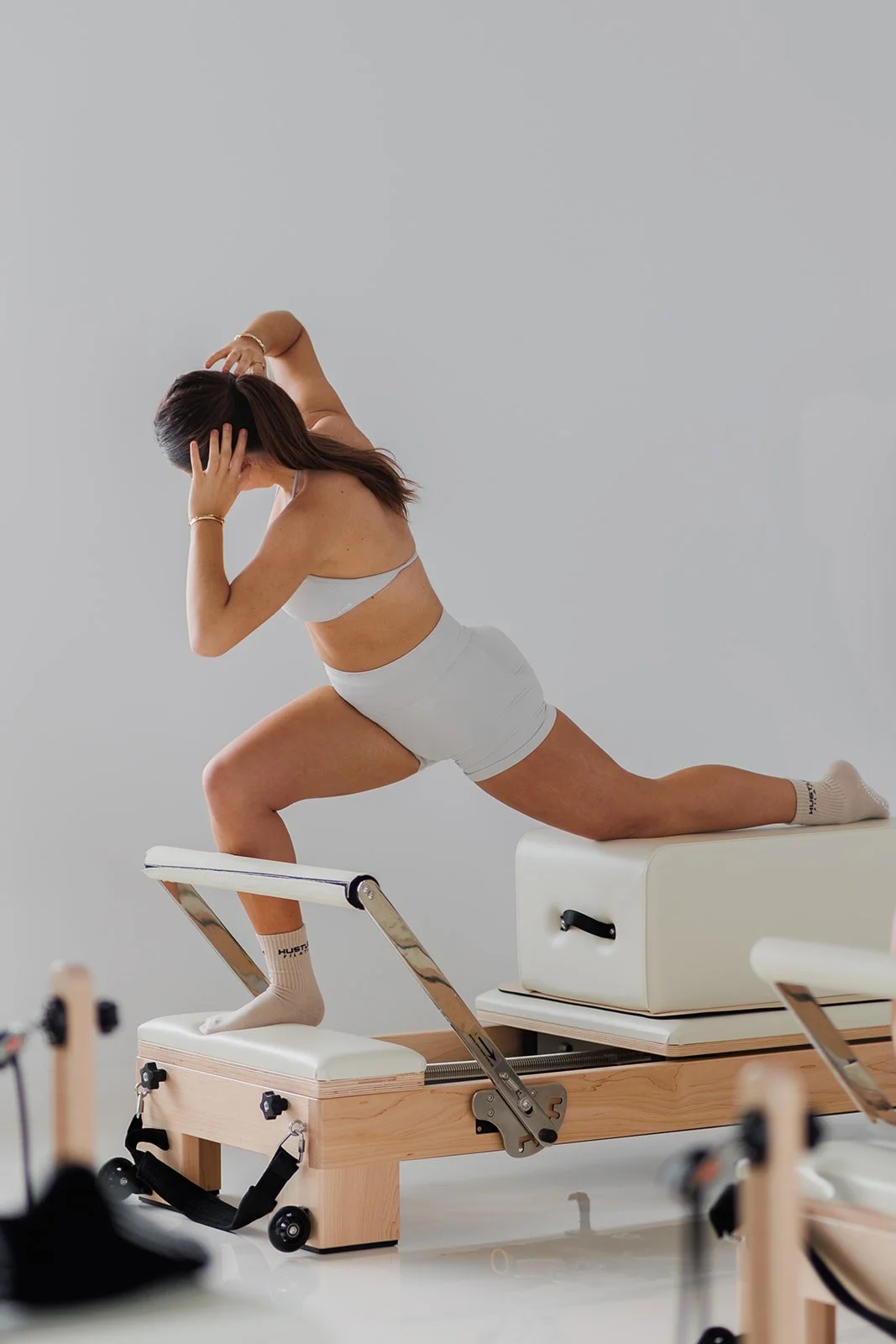 A woman practicing Pilates on a reformer machine, wearing activewear, with her hand on her head and knee on the padded surface.
