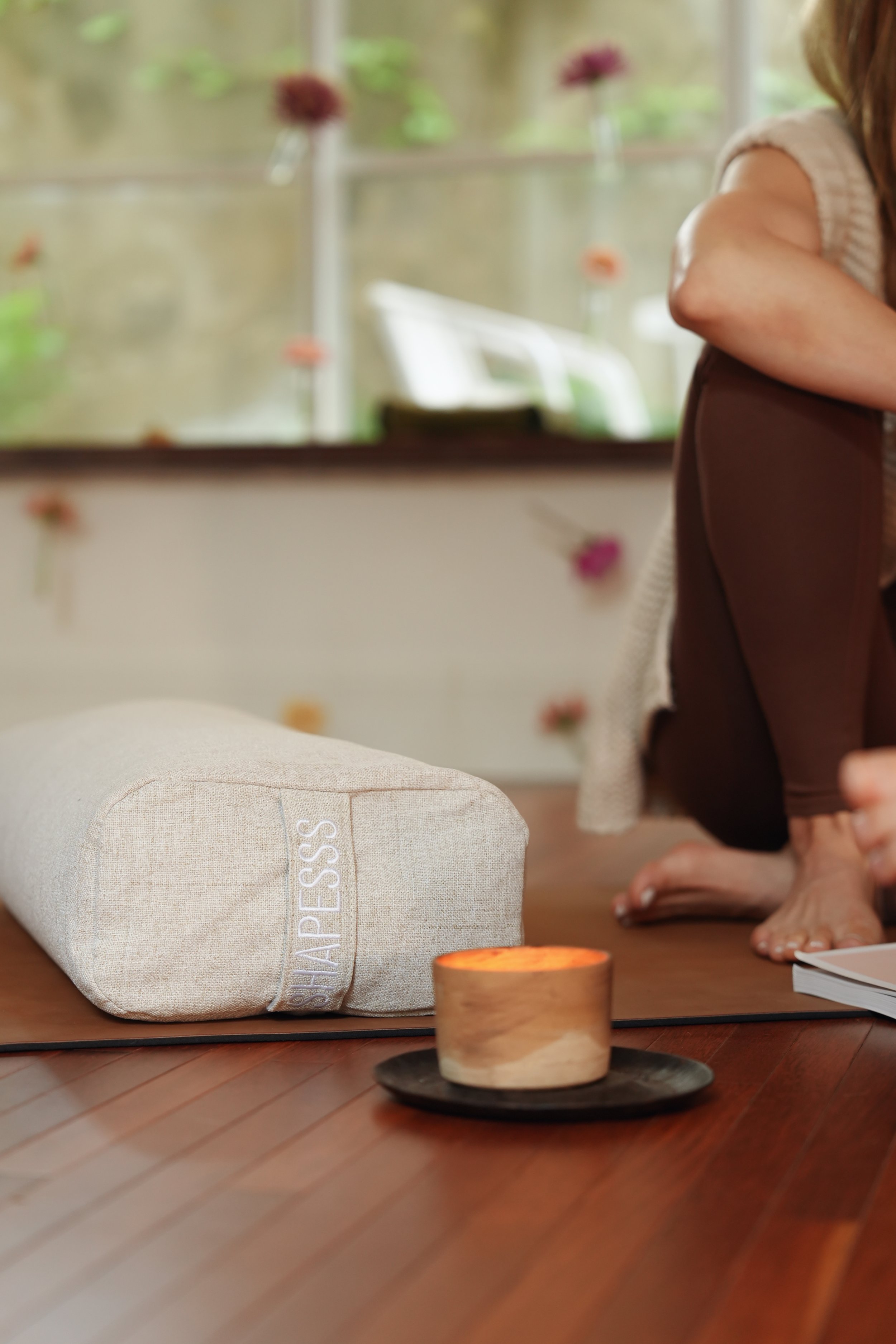 Close-up of a beige fabric wellness or massage pillow with the text 'HAPPINESS' embroidered on it, placed on a wooden floor in a room with a large window and some decorative objects, including a candle on a dark plate and a person practicing yoga or meditation nearby.