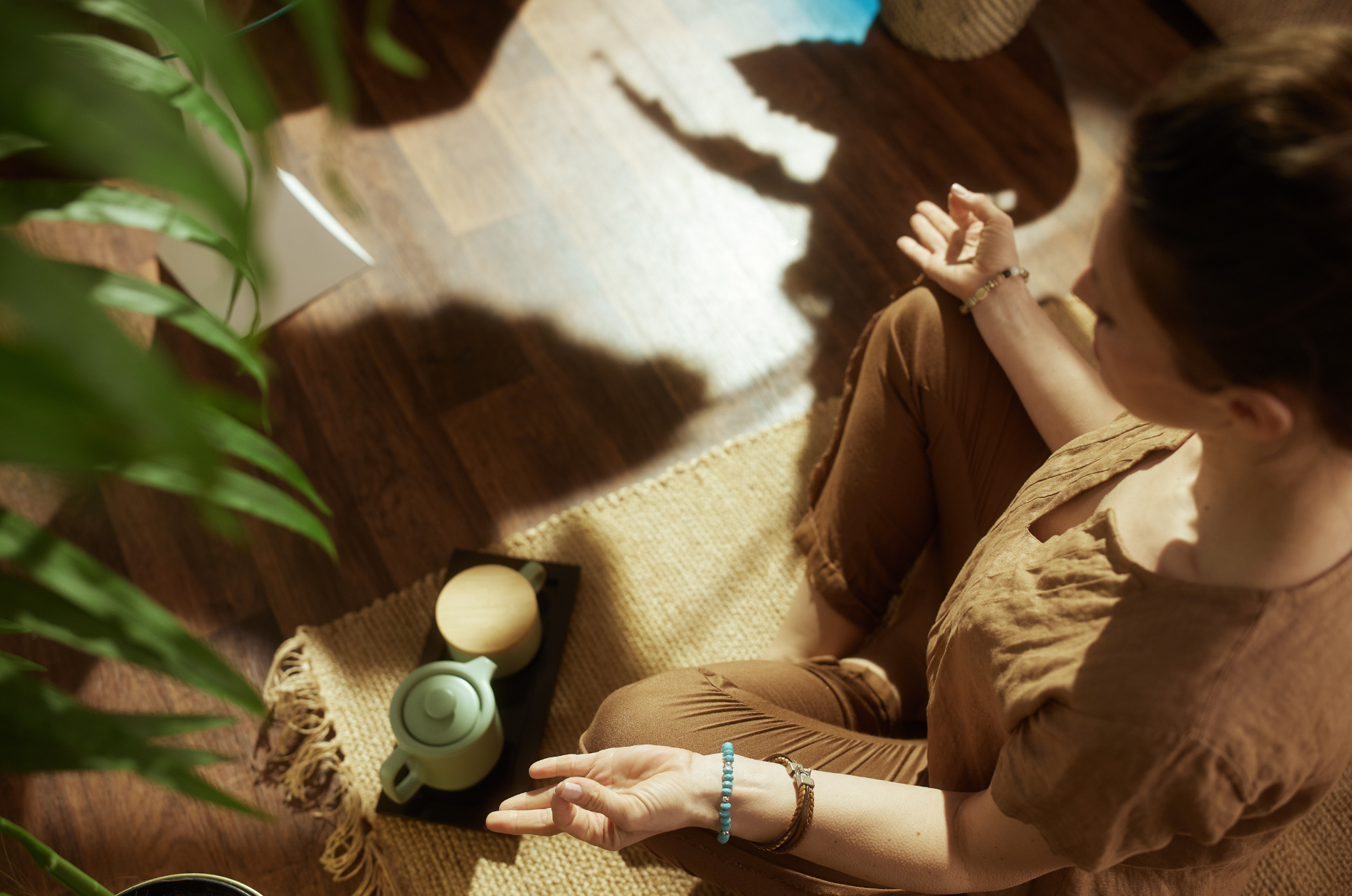 Woman sitting cross-legged on a natural fiber rug on a wooden floor, with a potted plant to her left. She is wearing brown clothing and jewelry, and is making a peace sign with her right hand. On the floor in front of her, there are a teapot and a matching cup.