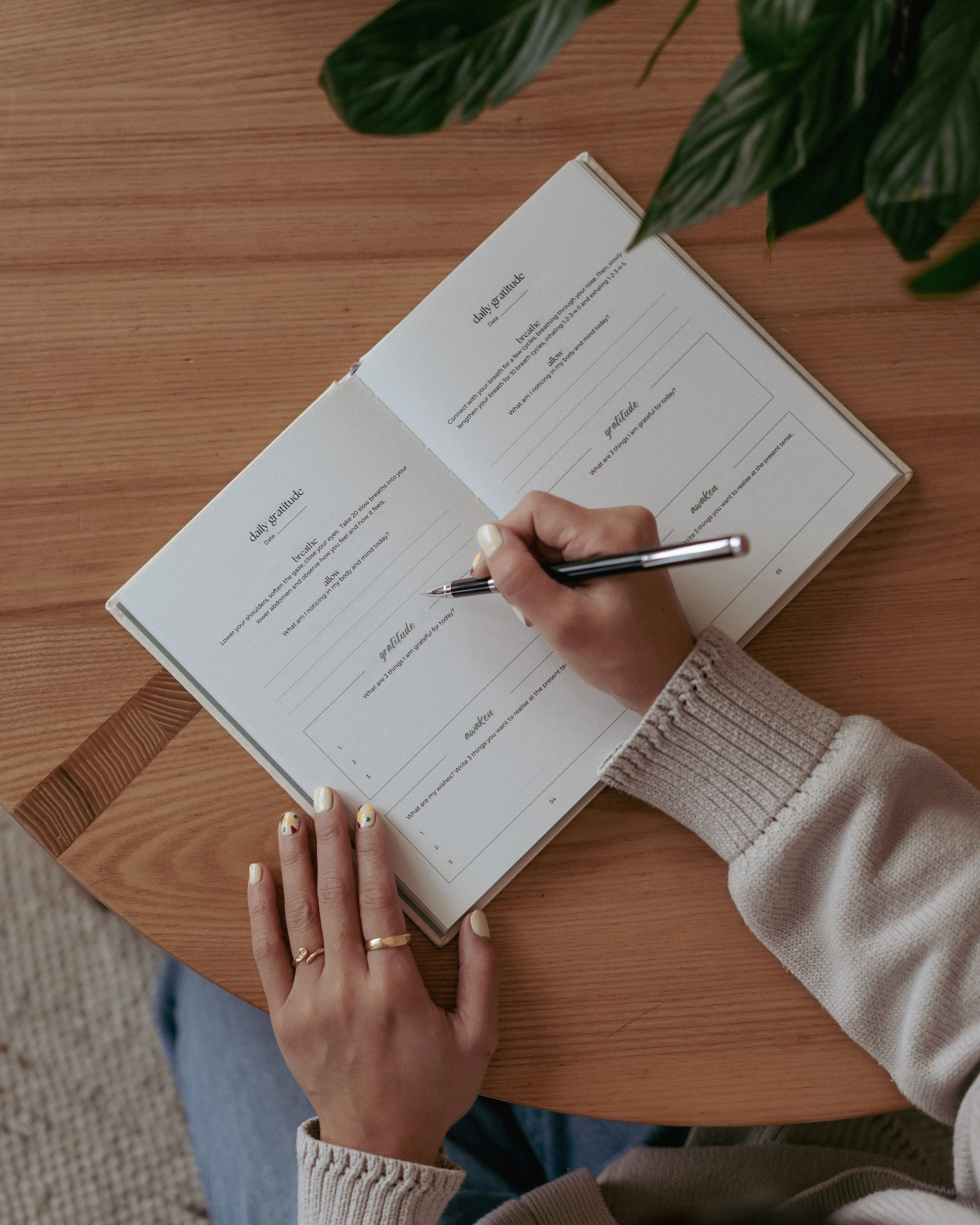 A person writing in a journal on a wooden table, surrounded by greenery, with decorated nails and wearing a beige sweater.
