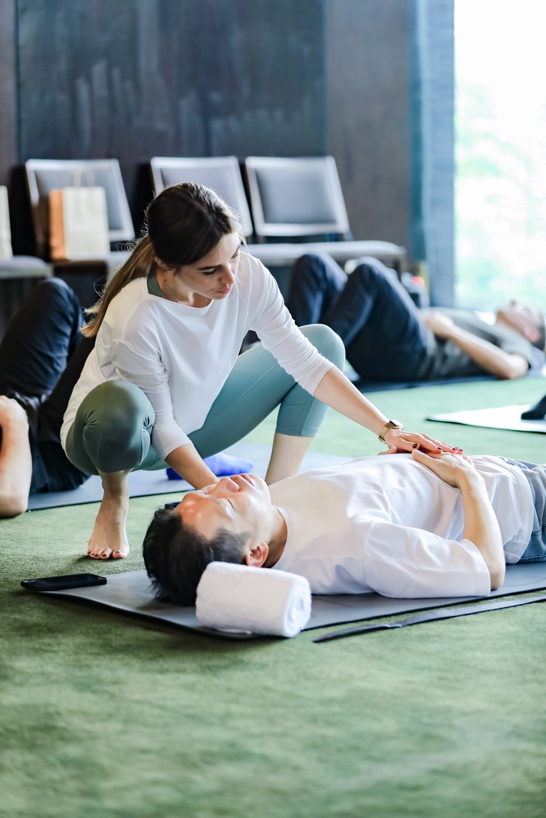 A woman practicing CPR on a man lying on a mat in a training class, with other people and chairs in the background.
