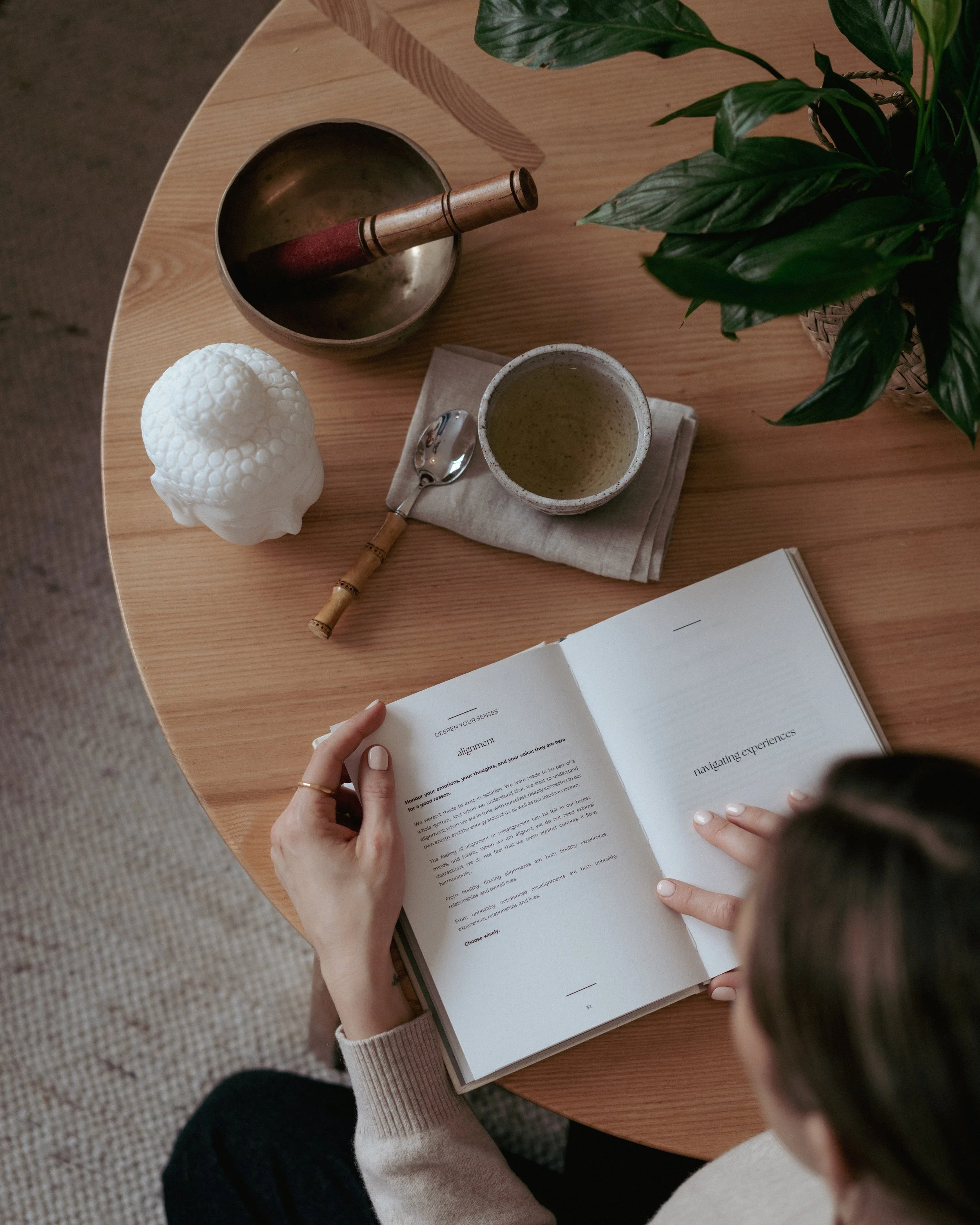 A person reading a book featuring the words 'Deepen Your Senses' and 'alignment' on a page, sitting at a round wooden table. On the table are a small spoon with a wooden handle, a ceramic cup with a hot beverage, a decorative white object, a pouch, and a green leafy plant in a basket.