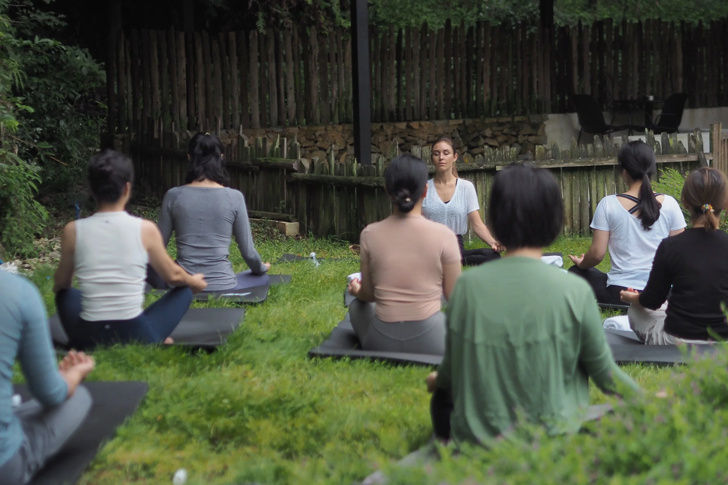 A group of people practicing yoga outdoors in a forested area, sitting in a circle on yoga mats with a woman leading the session.