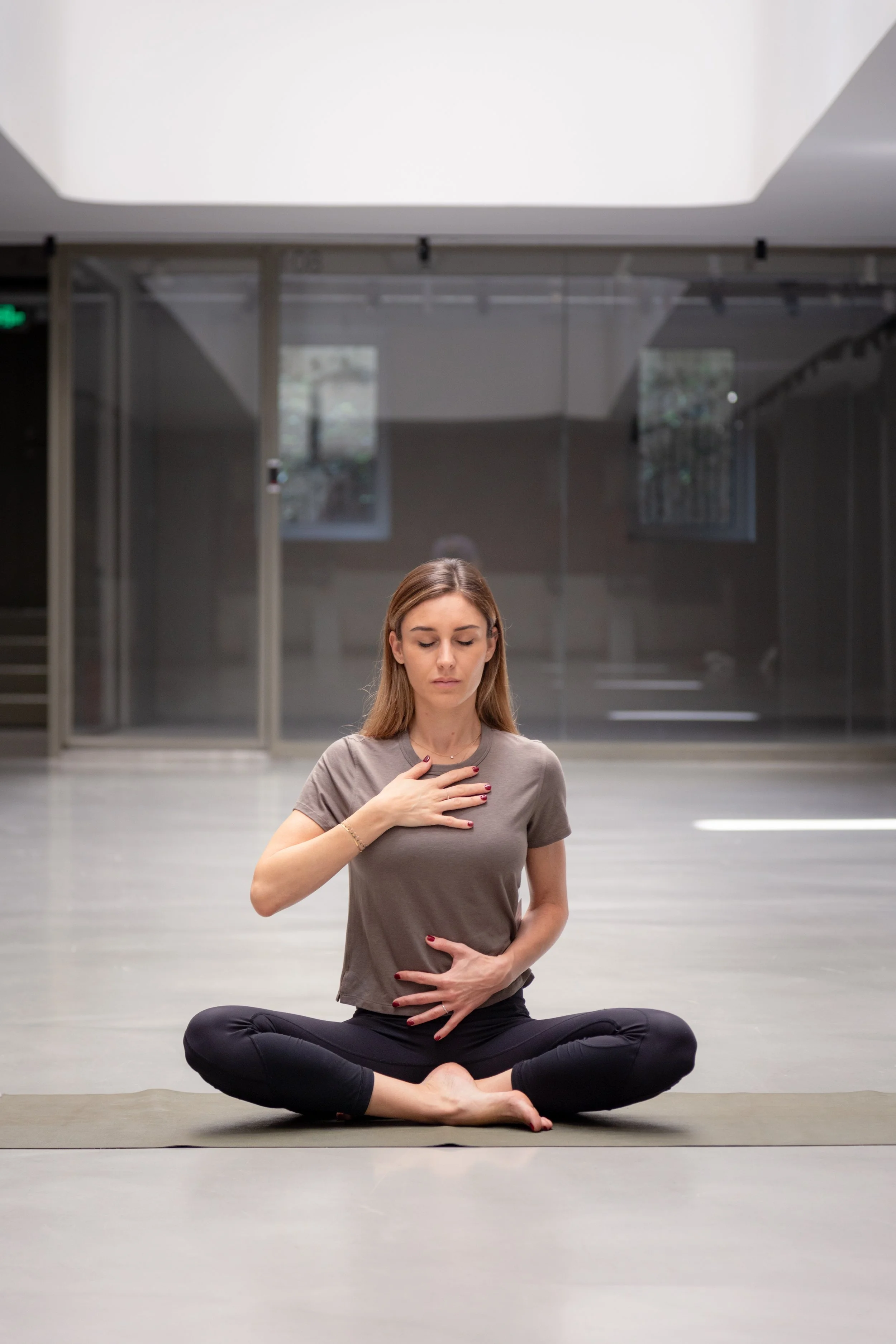 A woman practicing yoga indoors, seated cross-legged on a mat, with one hand on her chest and the other on her stomach, eyes closed, in a meditative pose.