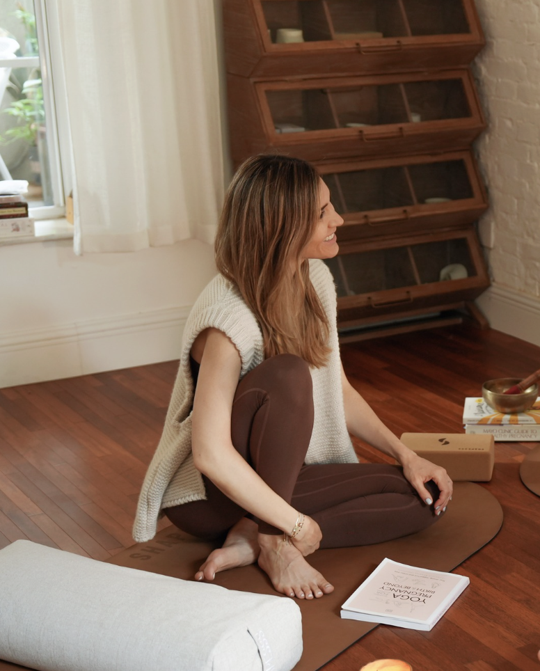 A woman with long, wavy brown hair sitting cross-legged on a mat, smiling, in a cozy room with wooden floors and a white brick wall.