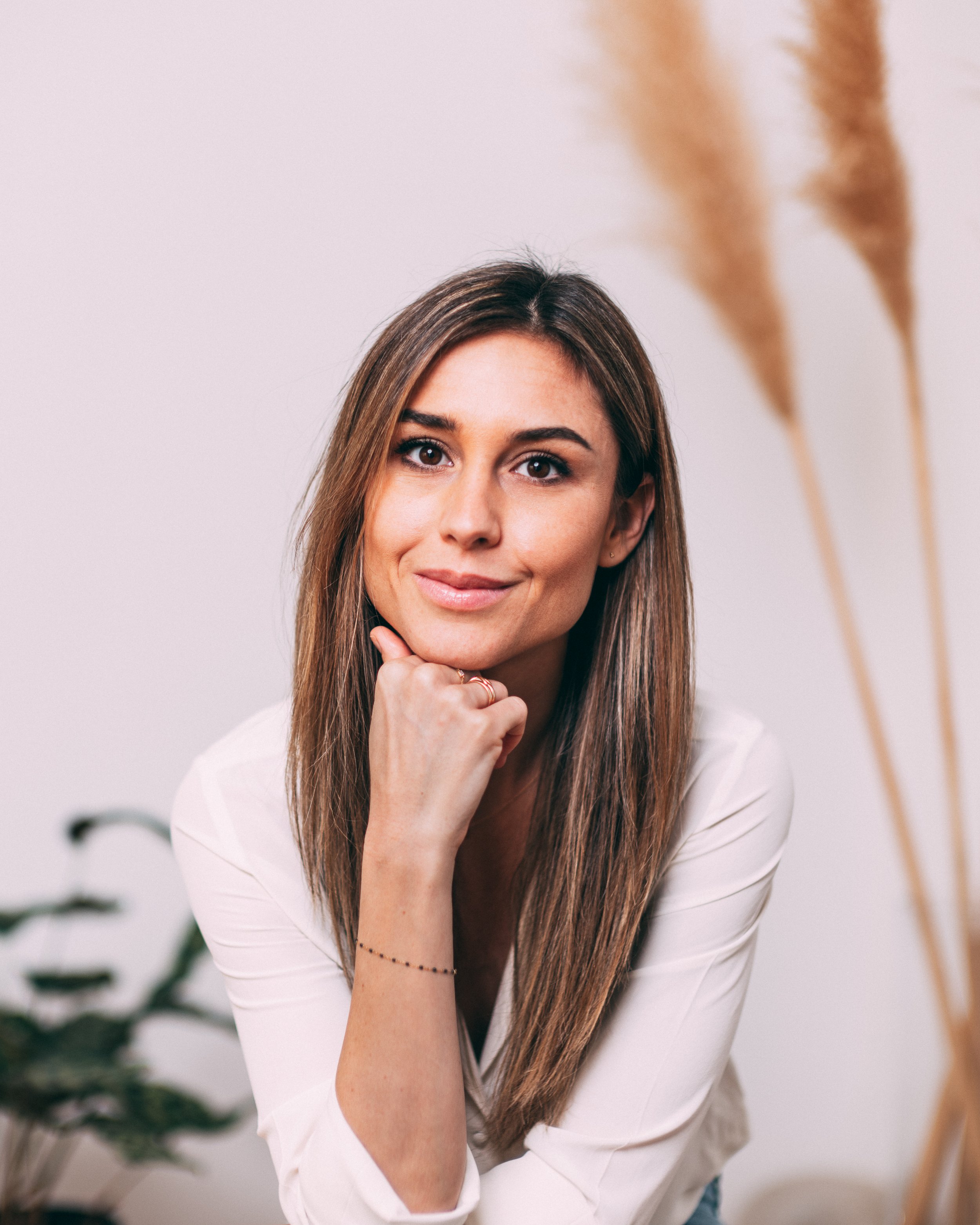 Close-up of a woman with long brown hair, smiling subtly, wearing a white top and jewelry, indoors with a neutral background and some green plants.