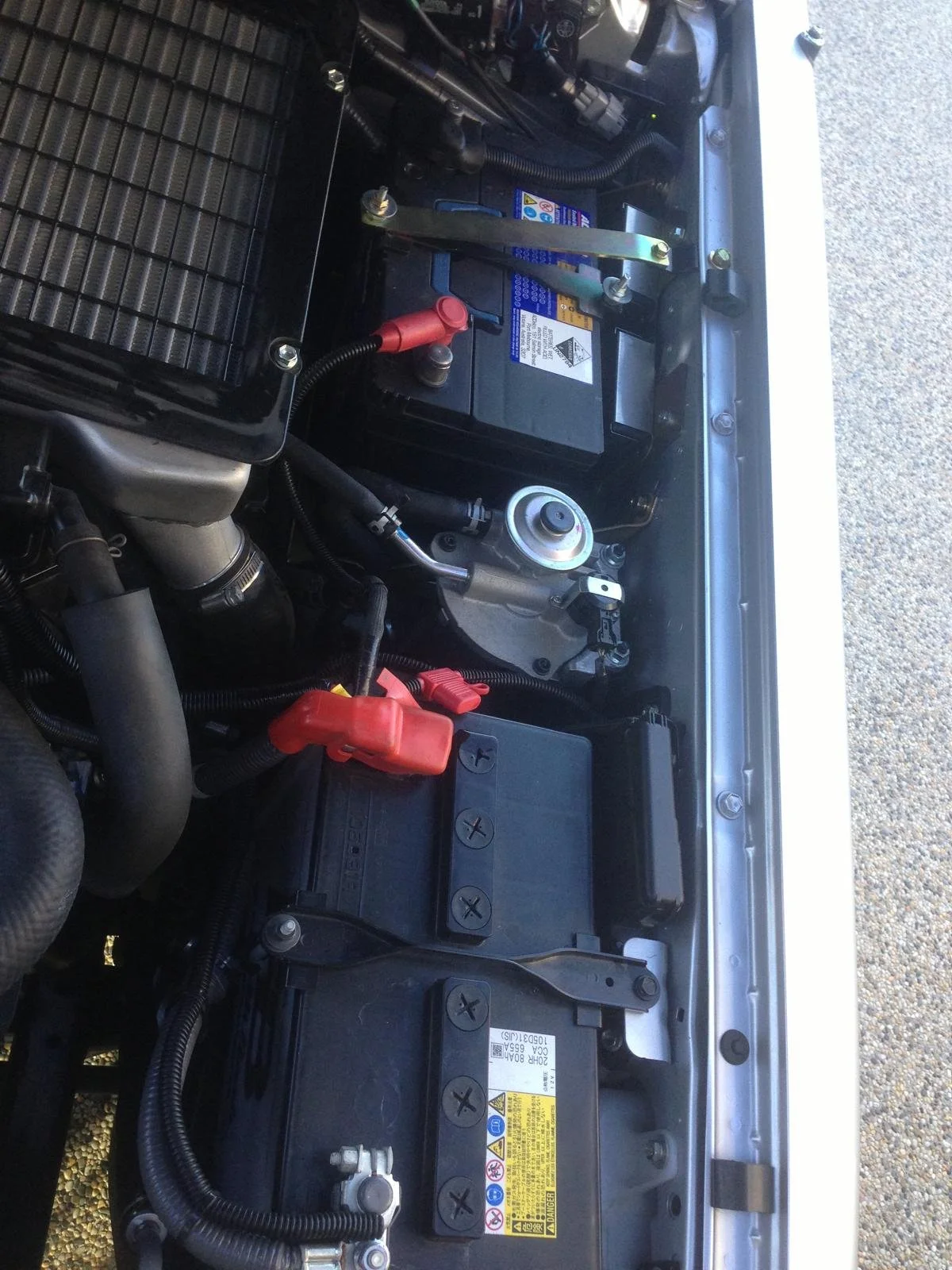 Close-up of vehicle engine compartment showing battery, radiator, and various parts, with a gravel surface outside the engine bay.