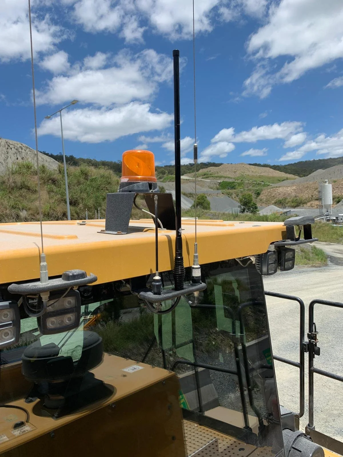 Close-up of the roof of a construction vehicle with orange warning light, antennas, and equipment, with a landscape of hills and partly cloudy sky in the background.