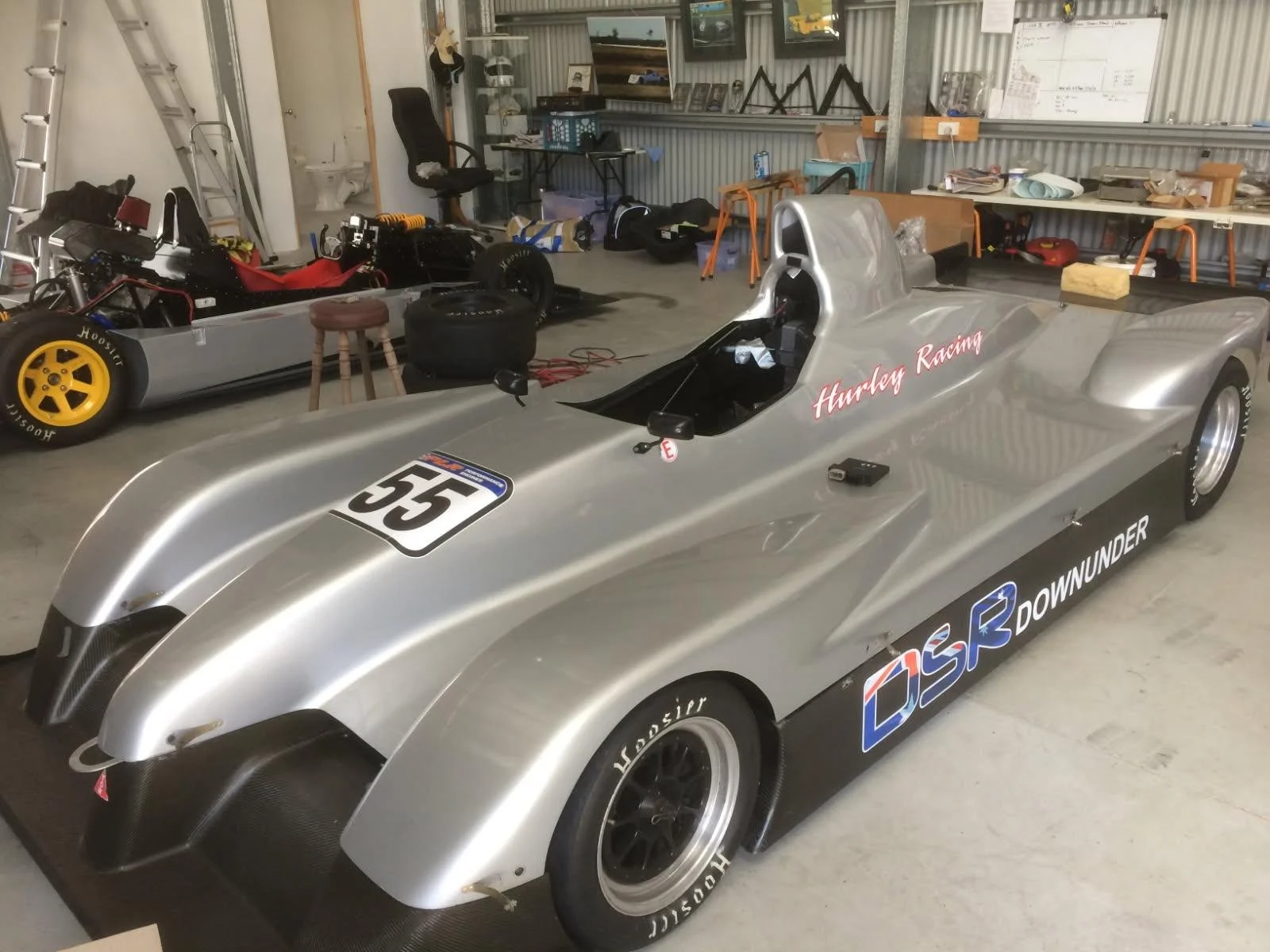 A silver race car with the number 55 and sponsor logos, parked in a garage with tools and equipment on shelves in the background.