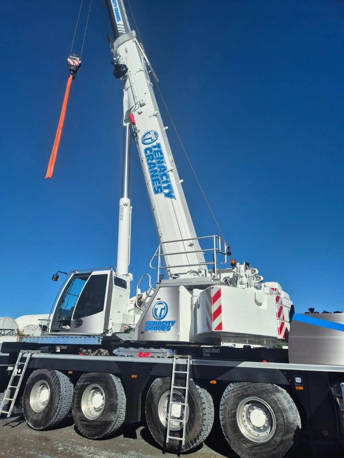 A large white crane with the logo 'Tenacity Cranes' and 'Heavy Cranes' on the boom, standing on a black flatbed trailer, with a clear blue sky in the background.