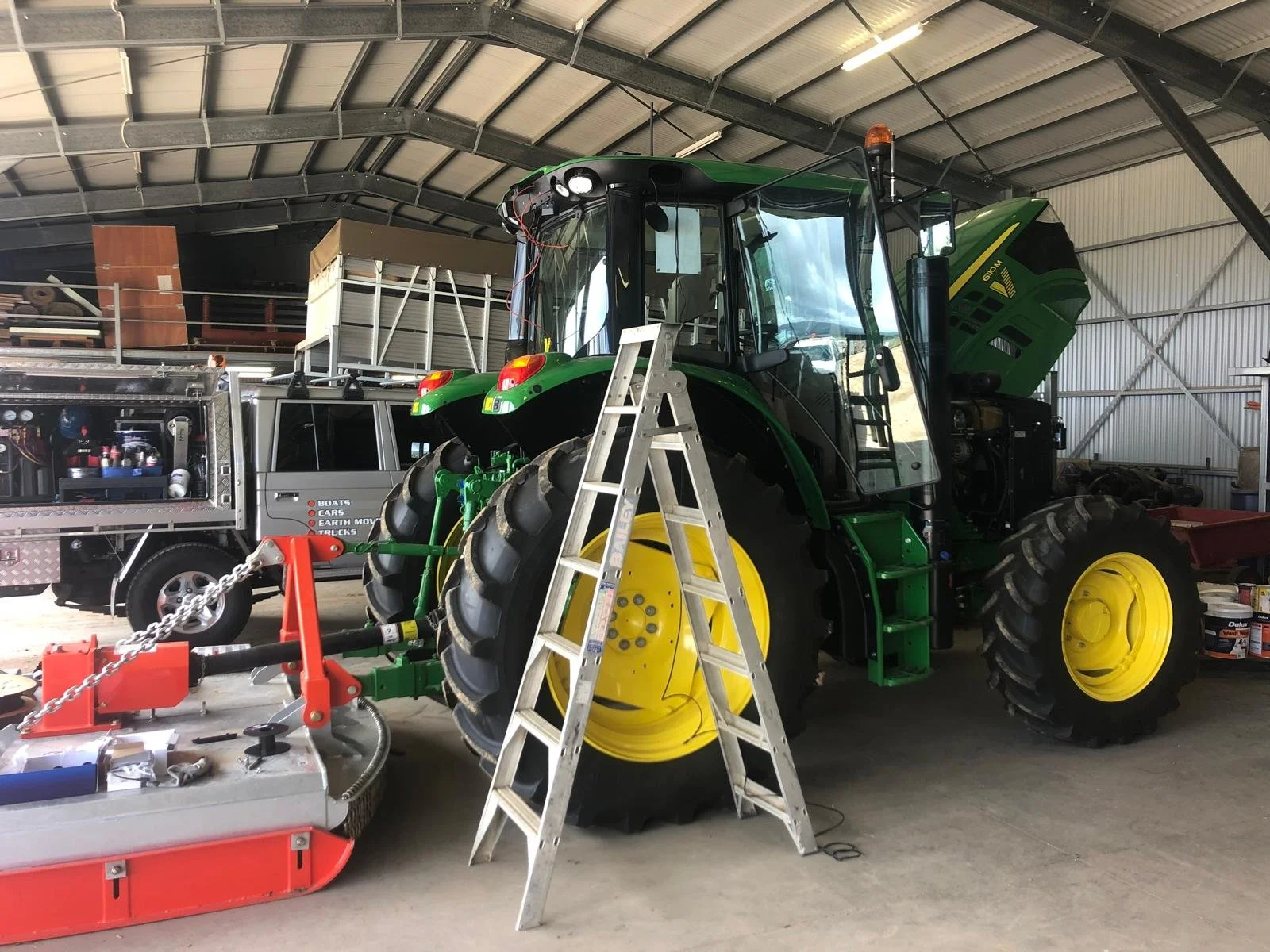 A green and yellow tractor inside a large garage or workshop, with a silver ladder in front and various tools and equipment around.