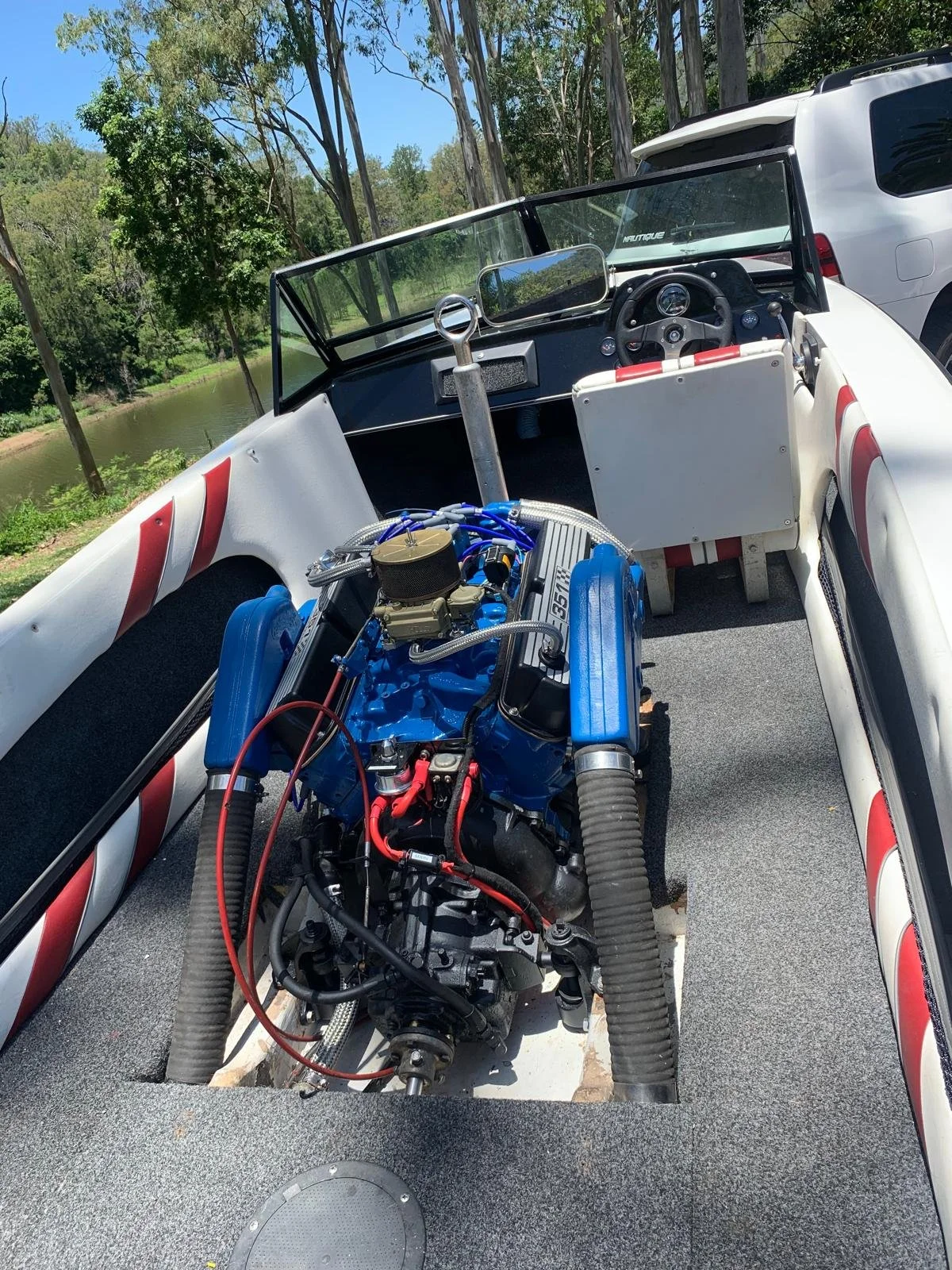 Race car with exposed engine in a boat, parked outdoors near trees and water.