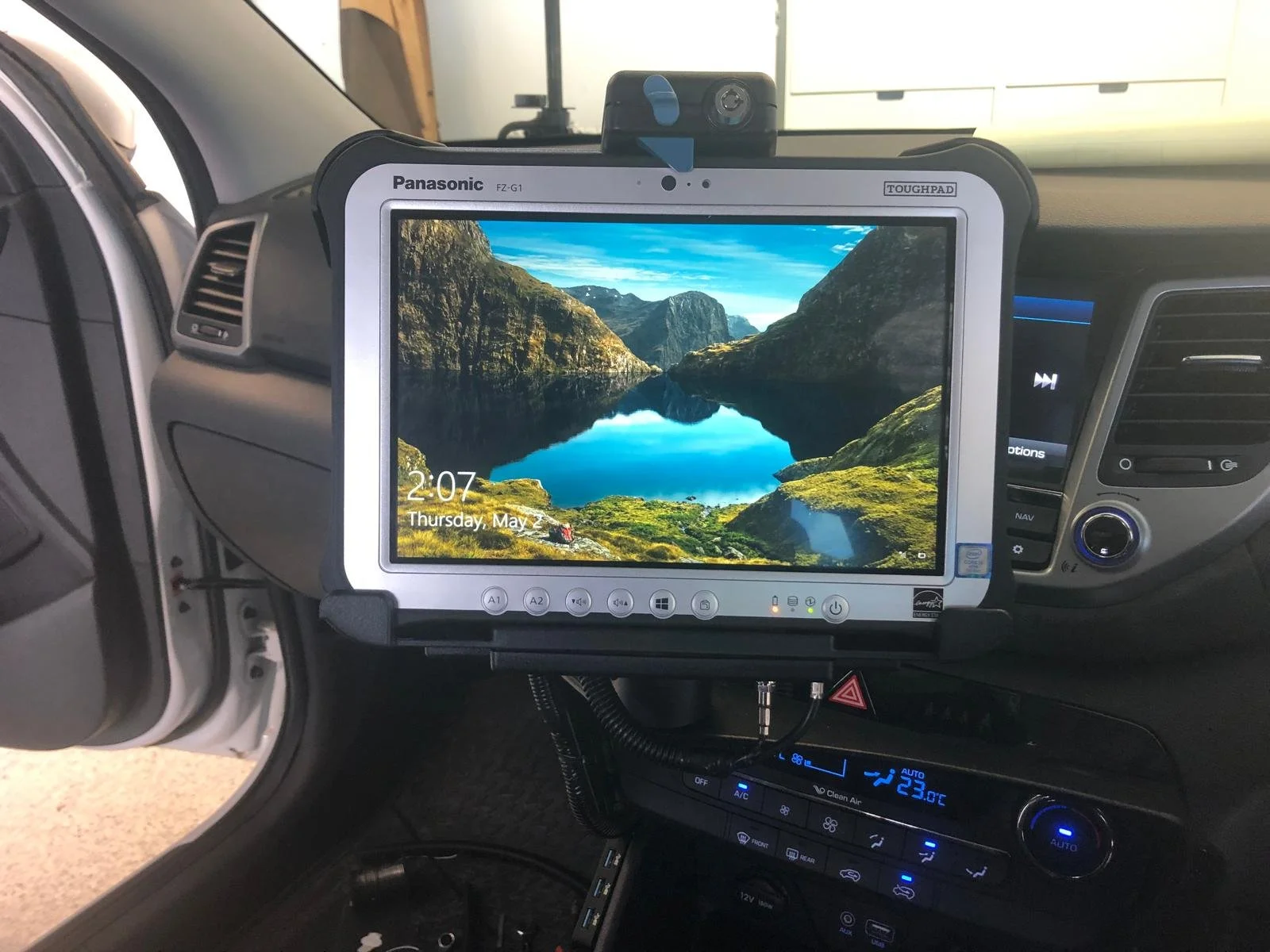 A dashboard inside a vehicle showing a mounted tablet displaying a scenic landscape of mountains and a lake. The tablet is branded Panasonic and is secured to the dashboard with a tough pad frame. The time and date on the tablet read 2:07 and Thursday, May 2.