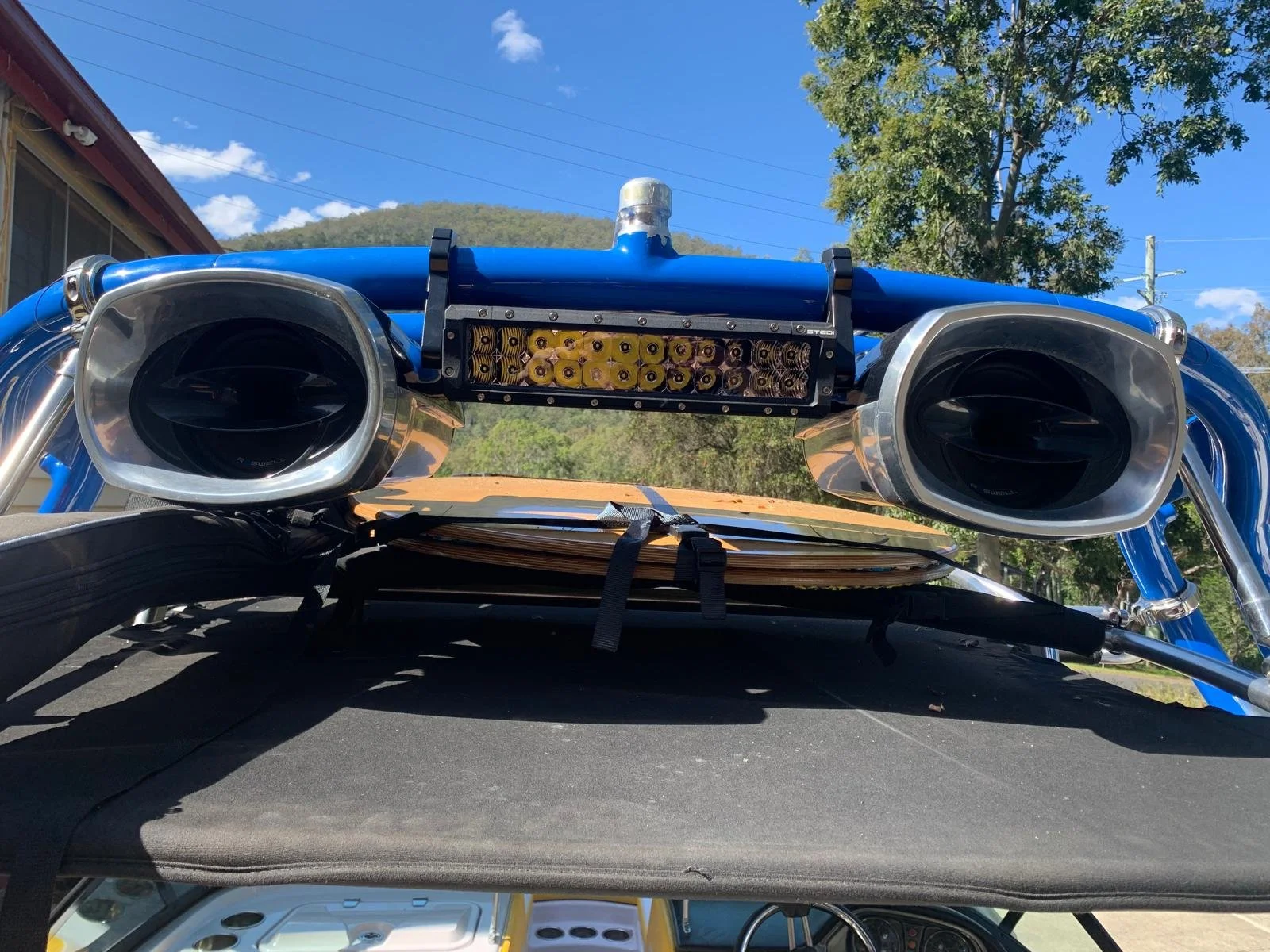 Close-up of a telescope mounted on a vehicle, with blue and silver metal components, black adjustment straps, and a yellow object underneath, against a bright blue sky with clouds and trees.