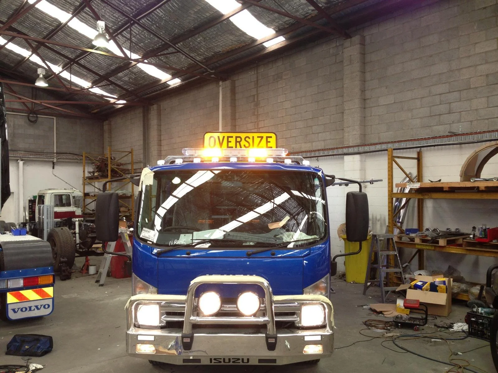 Blue construction or utility vehicle with an oversized sign on top, inside a workshop or warehouse with tools and equipment around.