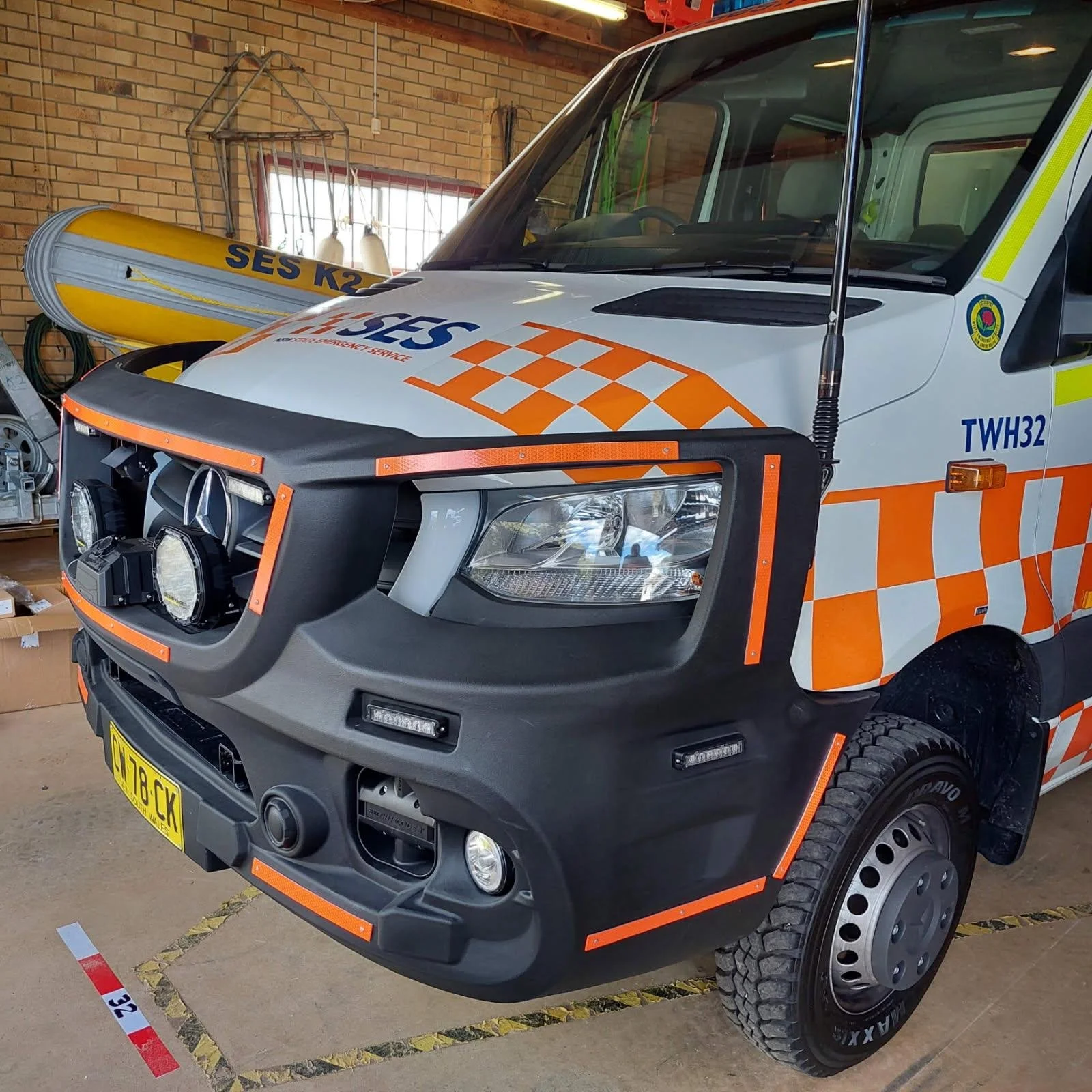 An emergency services vehicle with orange and white checkered patterns, a Mercedes-Benz logo on the front grille, yellow and blue markings, and equipment stored in the garage.