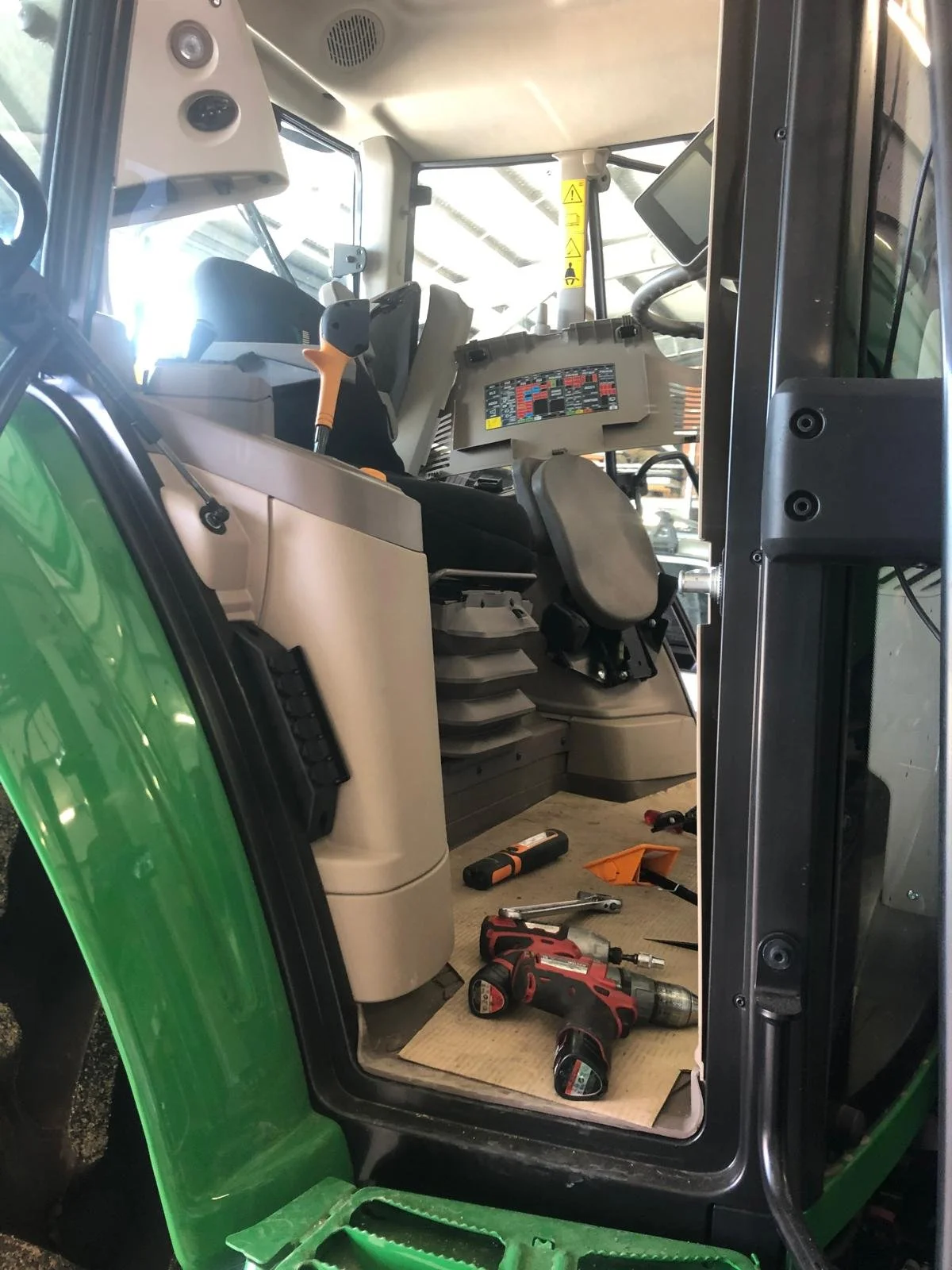 Inside the cab of a green tractor, showing the driver's seat, control panel, and various tools and equipment on the floor, including a cordless drill, screwdrivers, and a small toolbox.