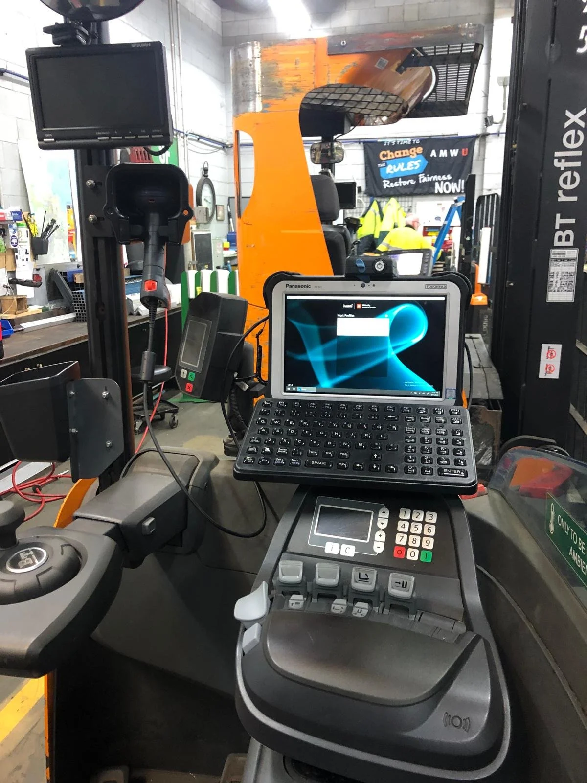 Interior of a forklift operator's cabin showing a computer screen, control panels, and forklift controls with a forklift lifting a pallet in a warehouse.